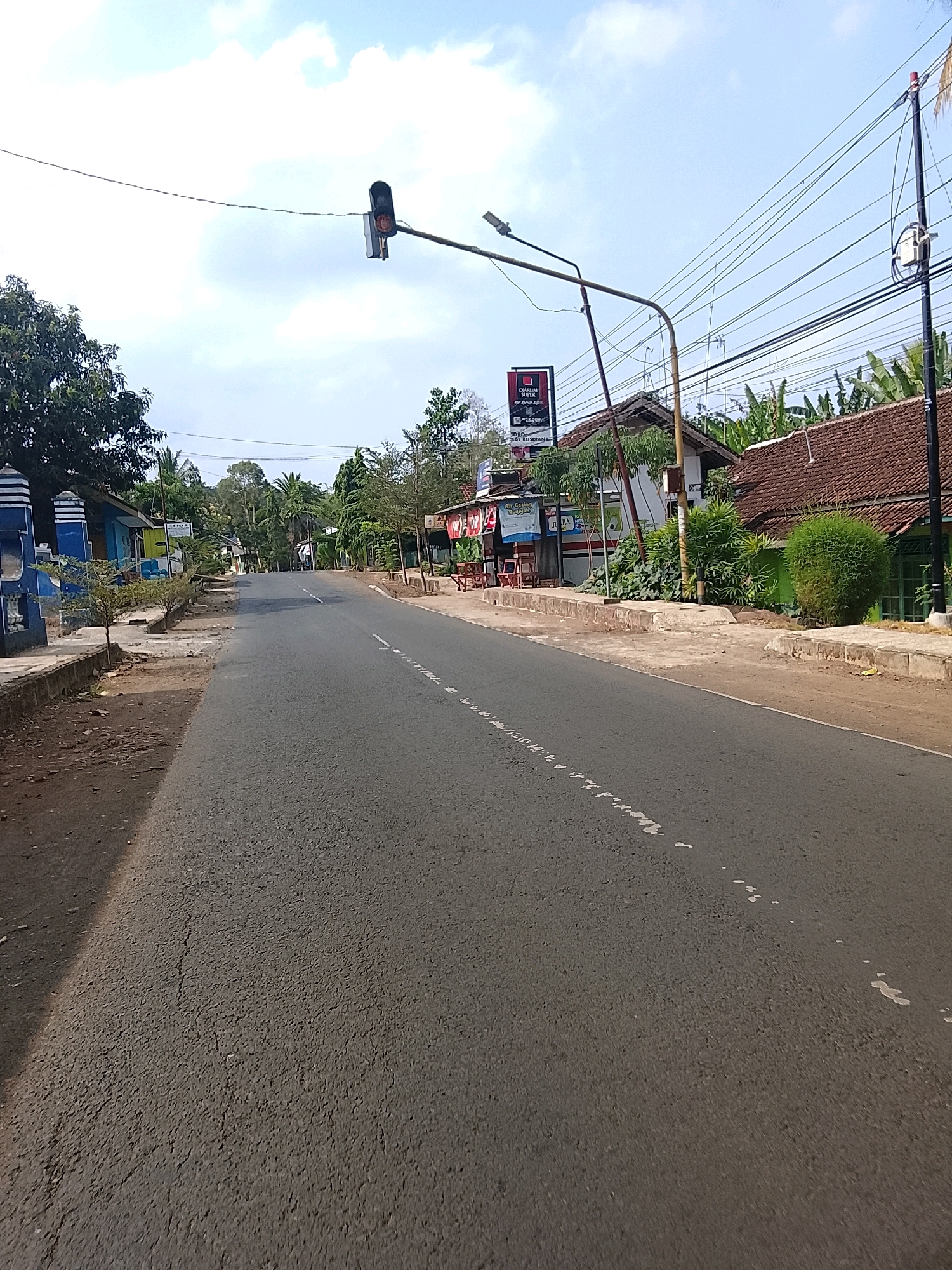 The image captures a street scene in Banjar, Indonesia, under a bright, partly cloudy sky. A two-lane asphalt road dominates the foreground, marked with intermittent white dashes on the right side. To the left, a sidewalk runs alongside the road, bordered by a low concrete wall and a dirt area. A blue and white decorative pillar stands on the far left.
Along the right side of the road, several buildings with tiled roofs are visible. One building features a red and white awning with chairs and tables arranged outside, suggesting a food stall or small eatery. A prominent sign with red lettering and a white silhouette is also present. Further down the street, lush green vegetation, including banana trees and various shrubs, grows. Utility poles with a dense network of electrical wires crisscross the sky.
A traffic light, with its red and yellow lights illuminated, hangs suspended over the road from a metal pole. In the distance, the road curves slightly and is lined with more trees and buildings, hinting at a moderately populated area within the city. The overall impression is a typical roadside scene in a tropical urban or semi-urban environment.