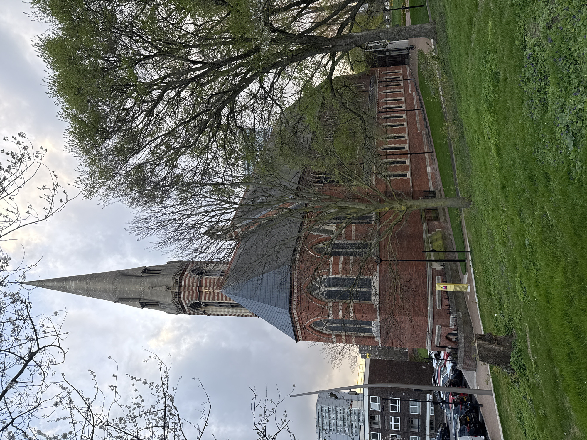 The image depicts a large brick church with a tall, pointed spire, partially obscured by trees with budding green leaves. The church has multiple arched windows and is bordered by a grassy area to the right. A sidewalk runs along the side of the church, and a yellow information kiosk is visible along the sidewalk. Behind the church and sidewalk, there is a row of parked cars and apartment buildings. The sky is overcast with patches of light. The location appears to be an urban area, possibly in London, United Kingdom, given the architectural style of the church and the surrounding buildings.