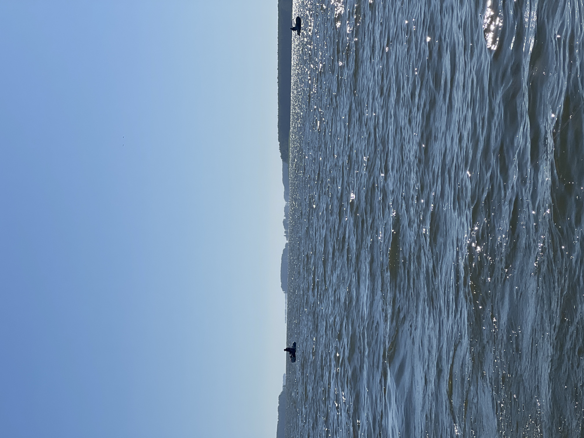 The image shows a wide expanse of dark blue water, with gentle waves creating shimmering highlights from the sun. In the distance, a tree-lined shore is visible on the right, and what appears to be land masses or islands stretch across the horizon. Two small boats are present on the water. The boat on the left contains a single silhouette of a person. The boat on the right also has a single silhouette of a person. The sky above is a clear, pale blue. The overall scene suggests an outdoor, possibly recreational, activity on a body of water. The location in Novopichugovo, Russia, implies this is likely a lake or river.