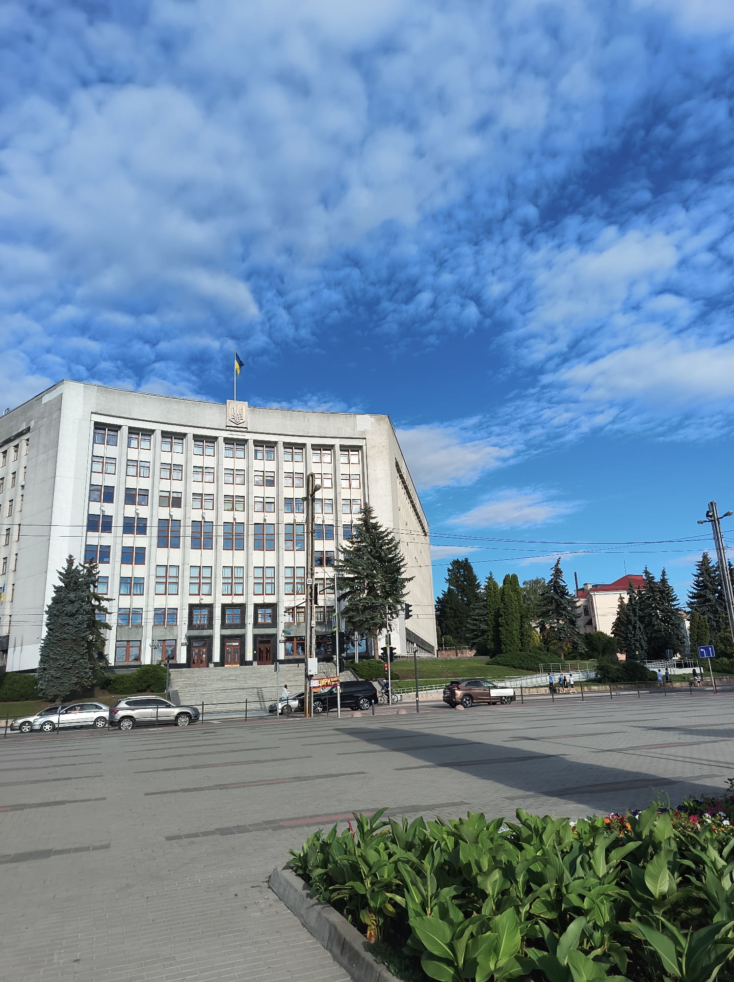 A wide, low-angle shot captures a large, brutalist-style building under a bright blue sky with scattered white clouds. A Ukrainian flag flies from a flagpole atop the building, which features numerous windows and a prominent coat of arms. Trees, including tall evergreens, are planted in front of and around the building. In the foreground, cars are parked on a wide, paved plaza with some landscaping visible at the bottom right, including green plants and flowers. The scene appears to be in a city square during the daytime, possibly late afternoon, as suggested by the long shadows cast across the plaza. There are a few people visible on the plaza, including some walking and others on bicycles, suggesting a normal day in a public space. The overall impression is one of civic architecture and urban life on a pleasant day.