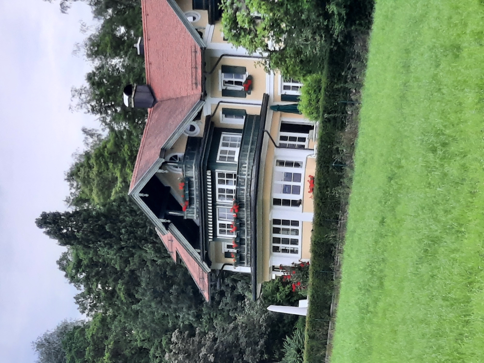 An aerial view of a large, multi-story house with a red tiled roof is captured from a slightly elevated perspective. The house is predominantly yellow with dark green trim and white window frames. It features balconies with flower boxes filled with red flowers on multiple levels. The house is set against a backdrop of dense green trees. A neatly trimmed hedge runs along the front of the property, separating it from a lush green lawn in the foreground. The sky is overcast. This scene is located in the city of Rosenheim, Germany.
