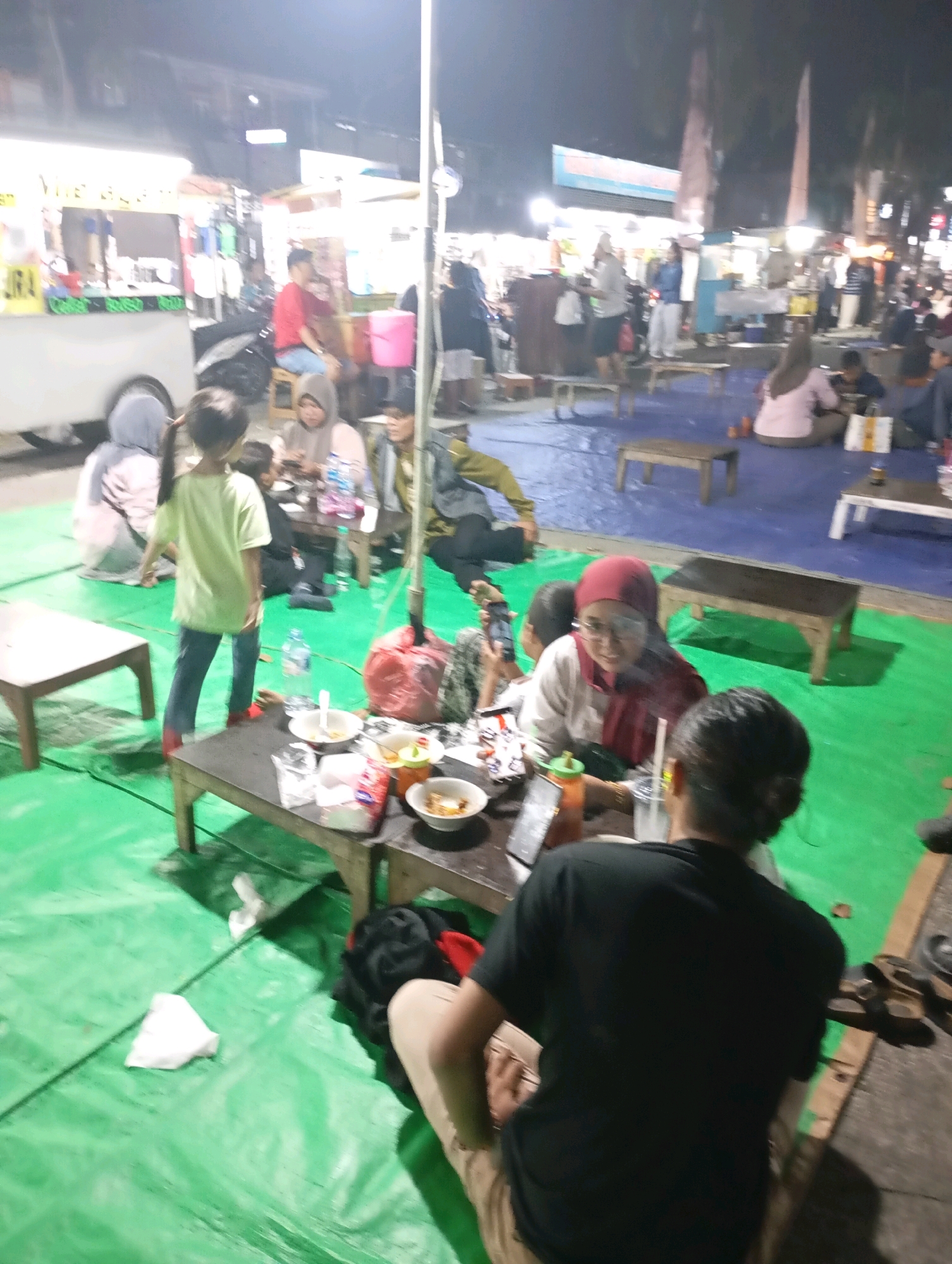 In Babelan, Indonesia, a group of people are gathered at night in an outdoor market setting, likely for dining. The scene is illuminated by artificial lights, creating a somewhat blurred and vibrant atmosphere.
In the foreground, a person wearing a black t-shirt and light-colored pants is seated facing away from the camera, appearing to interact with others at a low wooden table. The table is set with bowls, utensils, and a bottle of water, suggesting a meal is in progress. To the left of this table, a young girl in a light green t-shirt and blue jeans stands, facing the table, her hair in a ponytail.
Further back, several other individuals are seated around tables, some are eating or talking. One woman, wearing a red hijab and white top, is clearly visible and appears to be engaged in conversation. Another person in a hijab is seated next to her. In the mid-ground, a food stall with a white exterior and bright signage is present, with people standing and milling around it.
The ground is covered with green and blue tarps, indicating a temporary setup for the market. Wooden benches and low tables are scattered throughout the area. The overall impression is of a bustling night market with a focus on food and social gathering.
