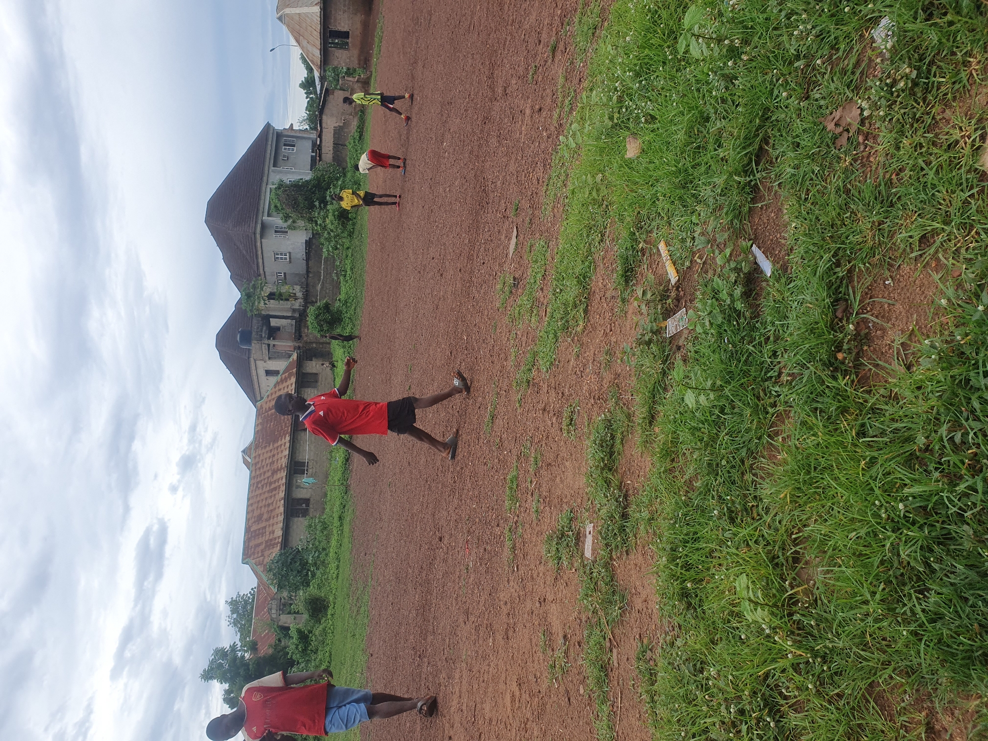 This image captures an outdoor scene in Idah, Nigeria, under an overcast sky. The ground consists primarily of reddish-brown dirt, with irregular patches of green grass, particularly in the foreground and lower right. Visible litter, including plastic wrappers and small discarded items, is present within the grassy areas.
Six male individuals are discernible within the frame. In the left foreground, a young male wearing a red and beige jersey with "ARSENAL" written across the chest, and blue shorts, walks towards the left. In the central mid-ground, another young male, wearing a red jersey, black shorts, and sandals, walks towards the right. Further into the mid-ground, three more males are present: one in a yellow jersey and black shorts stands facing the left; one in a white top and red shorts is in a stooping position, facing the ground; and one in a green jersey and black shorts stands with his back partially to the viewer, facing right. A sixth individual, dressed in dark clothing, is partially visible in the far left background, near the edge of the field.
The background features a residential area with various structures and vegetation. To the left, several older buildings with rusted corrugated metal roofs and weathered, unpainted walls are partially obscured by dense green trees and bushes. To the right, two larger, more modern-looking multi-story houses with dark, hipped roofs, light-colored facades, and multiple windows are prominent. These larger houses are surrounded by a low wall and some green foliage. A tall, dark streetlight pole is visible near the rightmost large house.