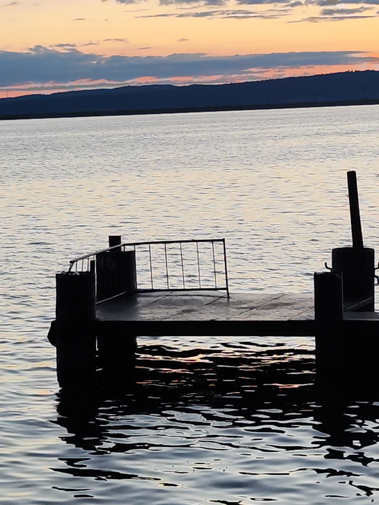 The image depicts a serene waterscape at either sunrise or sunset in Chaikovsky, Russia. A small, dark-silhouetted pier or dock extends into a calm body of water, likely a river or reservoir. No people are present in the scene.
The sky above is adorned with warm, soft hues of orange, pink, and light blue, suggesting the transitional light of dawn or dusk. Scattered clouds, ranging from wispy white to deeper blue-grey, are visible across the upper portion of the sky. A dark, low-lying landmass, possibly distant hills or mountains, forms the horizon line separating the water from the vibrant sky.
In the foreground, the pier is a prominent feature, rendered mostly in silhouette against the lighter water and sky. It comprises a flat platform supported by thick, dark posts or pillars. A metal railing with vertical bars is visible on one side of the platform. Notably, the number "16" is clearly marked in a lighter shade on the side of one of the dark, cylindrical structures or bollards near the railing. Another stout, dark post, possibly for mooring, stands towards the right side of the pier.
The water surface is gently rippled, reflecting the subtle colors of the sky and the dark forms of the pier structures. Closer to the pier, the water shows deeper shadows interspersed with faint glimmers of orange and yellow light. The overall atmosphere conveyed by the image is one of tranquility and peacefulness.