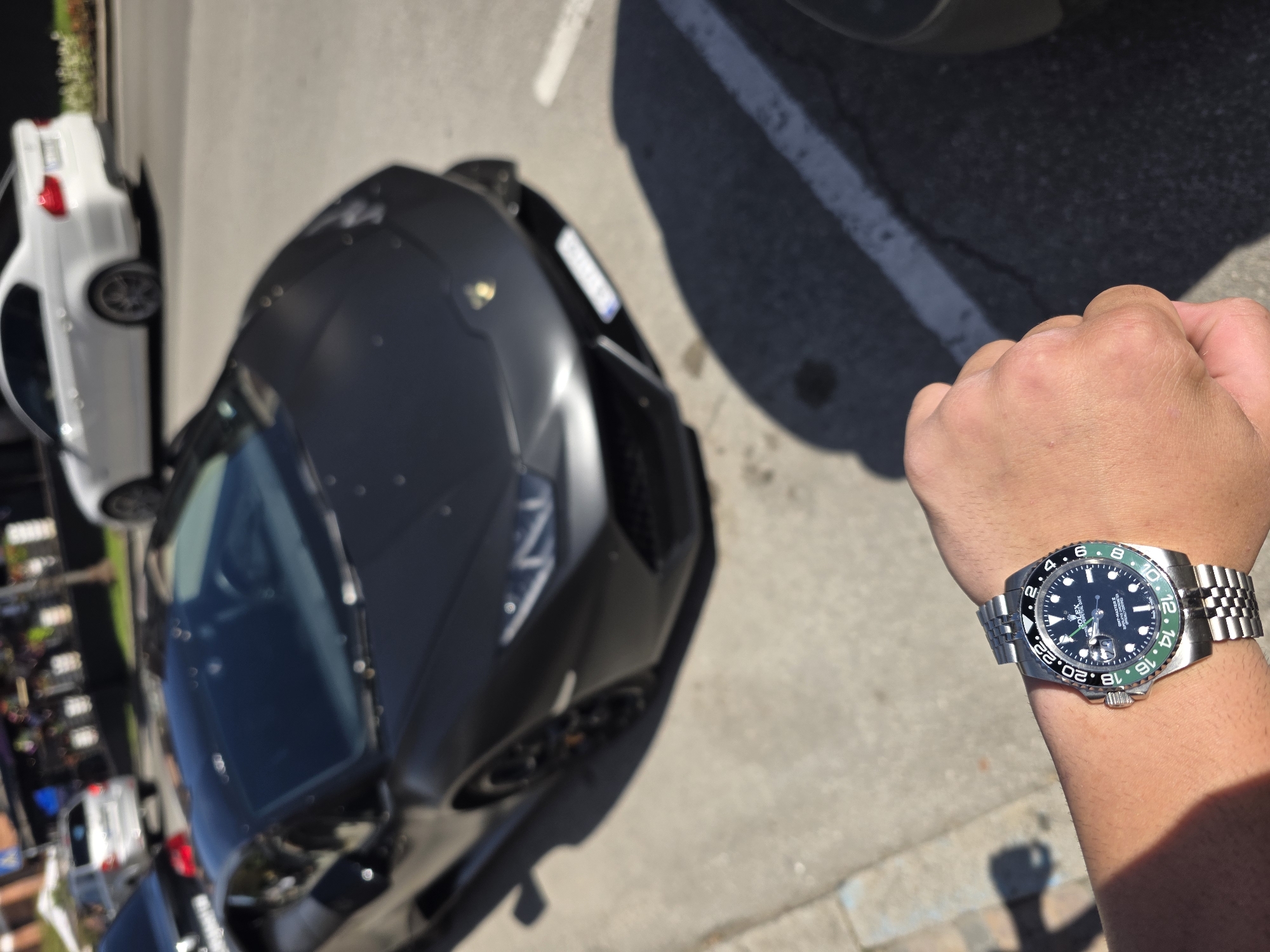 The image presents a wristwatch on a person's wrist, held out against the backdrop of a parking lot with cars. The watch is a Rolex GMT-Master II, with a black dial and a green and black bezel. The band is silver. Behind the wrist, there is a dark grey Lamborghini with a visible license plate obscured by shadow. To the left of the Lamborghini, there is a white vehicle. The background includes other vehicles and hints of greenery, suggesting an urban or suburban setting. The lighting indicates it is daytime with bright sunlight. The scene is captured from a high-angle, point-of-view shot, likely taken by the person wearing the watch. The location in Veliko Tarnovo, Bulgaria is not evident from the image.