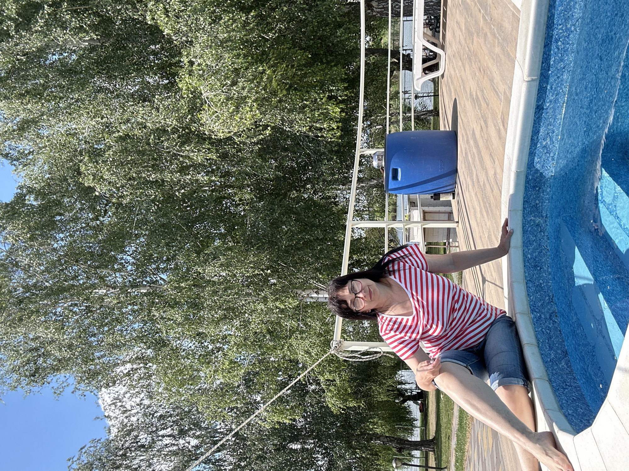 A woman with dark hair and glasses, wearing a red and white striped t-shirt and denim shorts, is sitting on the edge of a blue pool. She is on a wooden deck surrounded by trees and a white railing. In the background, a large blue barrel and white lounge chairs are visible. The scene appears to be outdoors, likely in a recreational area.