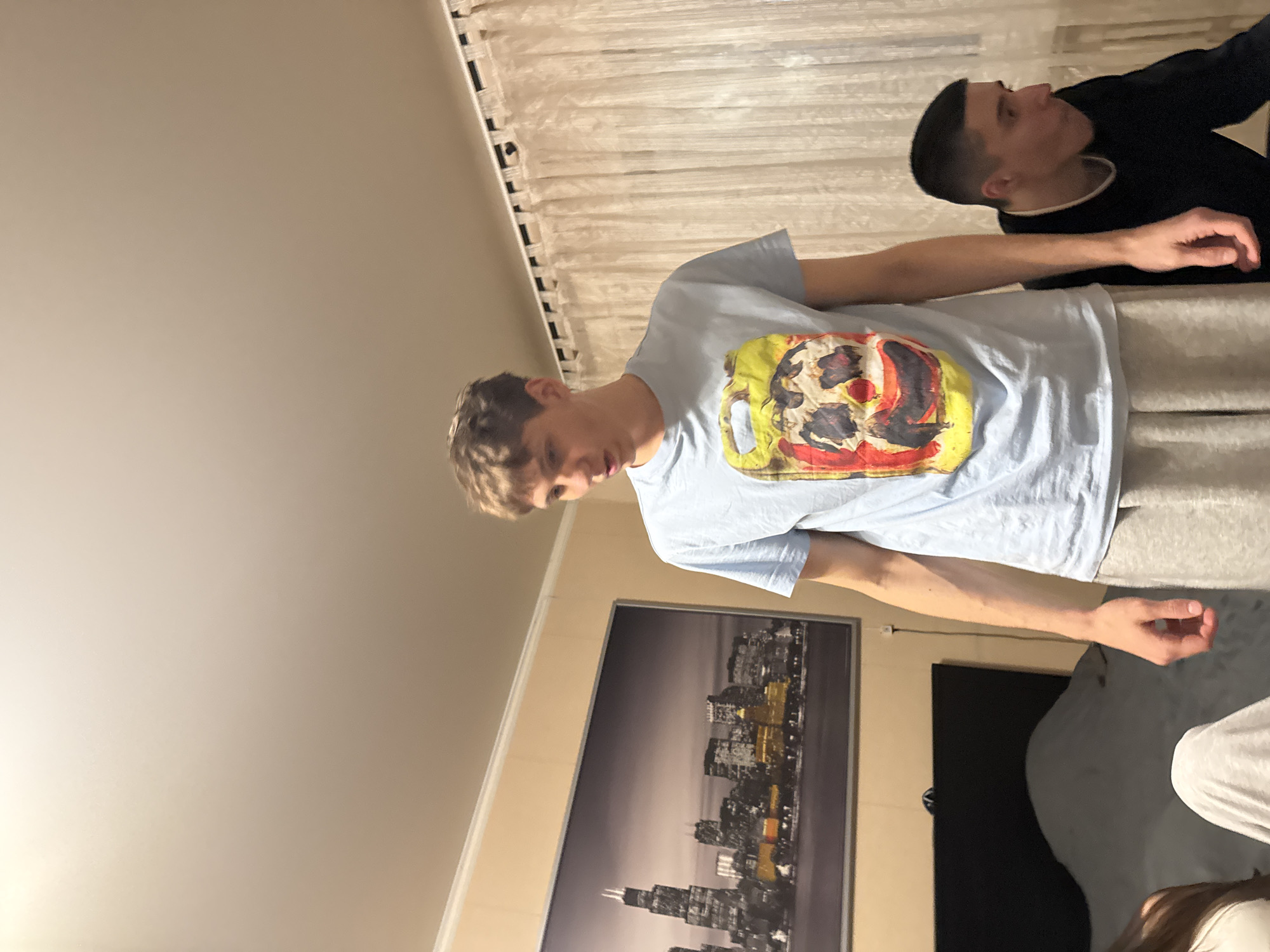 Two young men are in a room. One is wearing a light blue t-shirt with a graphic print and gray sweatpants. The other is wearing a black top with white lining at the neck. The room has light-colored walls and a white curtain. A picture depicting a skyline of a city hangs on the wall, possibly of Chicago. The location is not Kazan, Russia.