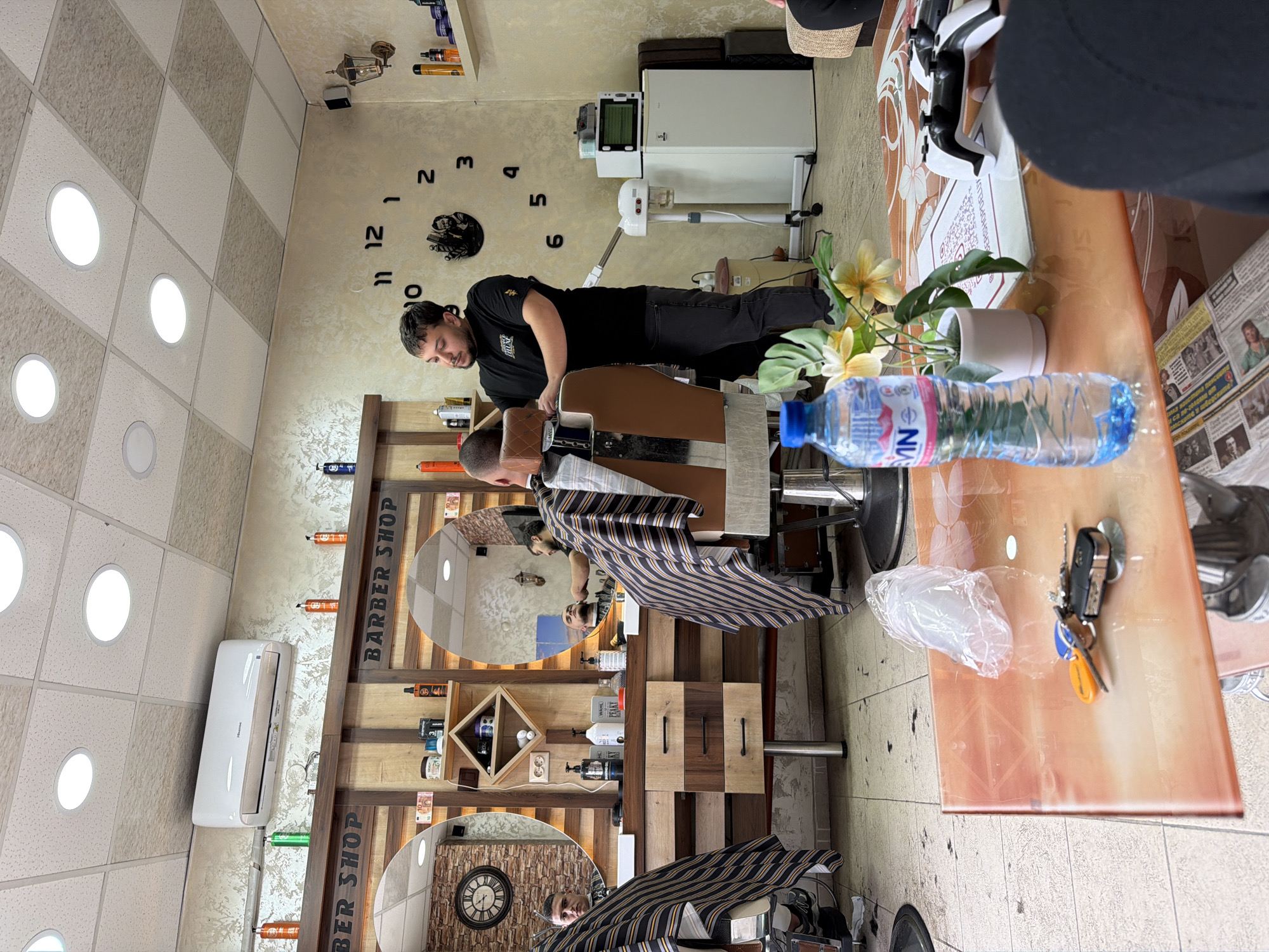 The interior of a barbershop, identifiable by the “BARBER SHOP” sign and barbershop-themed decor, shows a barber giving a customer a haircut. The barber, wearing a black t-shirt, is standing beside the customer, who is seated in a barber’s chair with a striped cape. The barbershop has a rustic, wooden theme with mirrors reflecting the scene. The wall decorations include shelves of barber supplies and clocks showing different times.
In the foreground, a table displays items like a water bottle, a potted plant, plastic bags, and a set of keys, adding a personal touch. The location is suggested to be in Panagyurishte, Bulgaria, based on the name "MN" and a name on the bottle, which may be a regional brand or identifier.