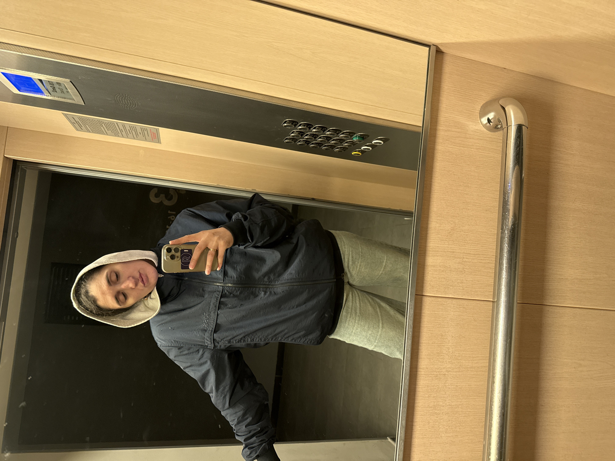 This media file captures a mirror selfie taken by a female individual inside an elevator, likely located in Tobolsk, Russia. The person, appearing to be in her late teens or twenties, has dark hair and closed eyes, suggesting a relaxed or posed expression. She is wearing a dark blue or black hooded jacket with a light-colored hood and light grey sweatpants. A ring is visible on her left ring finger. She holds a smartphone with a light gold/bronze case, positioned to take the photograph.
The elevator's interior features light-colored wooden paneling on the walls. Above the mirror, a dark metallic control panel is visible. It includes a blue digital display showing the number "3", indicating the elevator's current floor. Below the display, text reads "MAX. LOAD 8 PERS" and "630 KG". Multiple silver buttons are arranged on the panel, with some illuminated in green and yellow/gold. Another small metallic plaque with unreadable text is situated beneath these details.
To the right, a polished silver metallic handrail is attached to the wooden wall. The reflection in the mirror also shows a dark floor. On the person's jacket, the reversed numbers "2022" are clearly visible. A large "3" is reflected on the floor or a wall behind the person, along with what appears to be "jp" or "gr". The symbol on the phone case is indistinct. The scene is illuminated by the elevator's interior lighting.