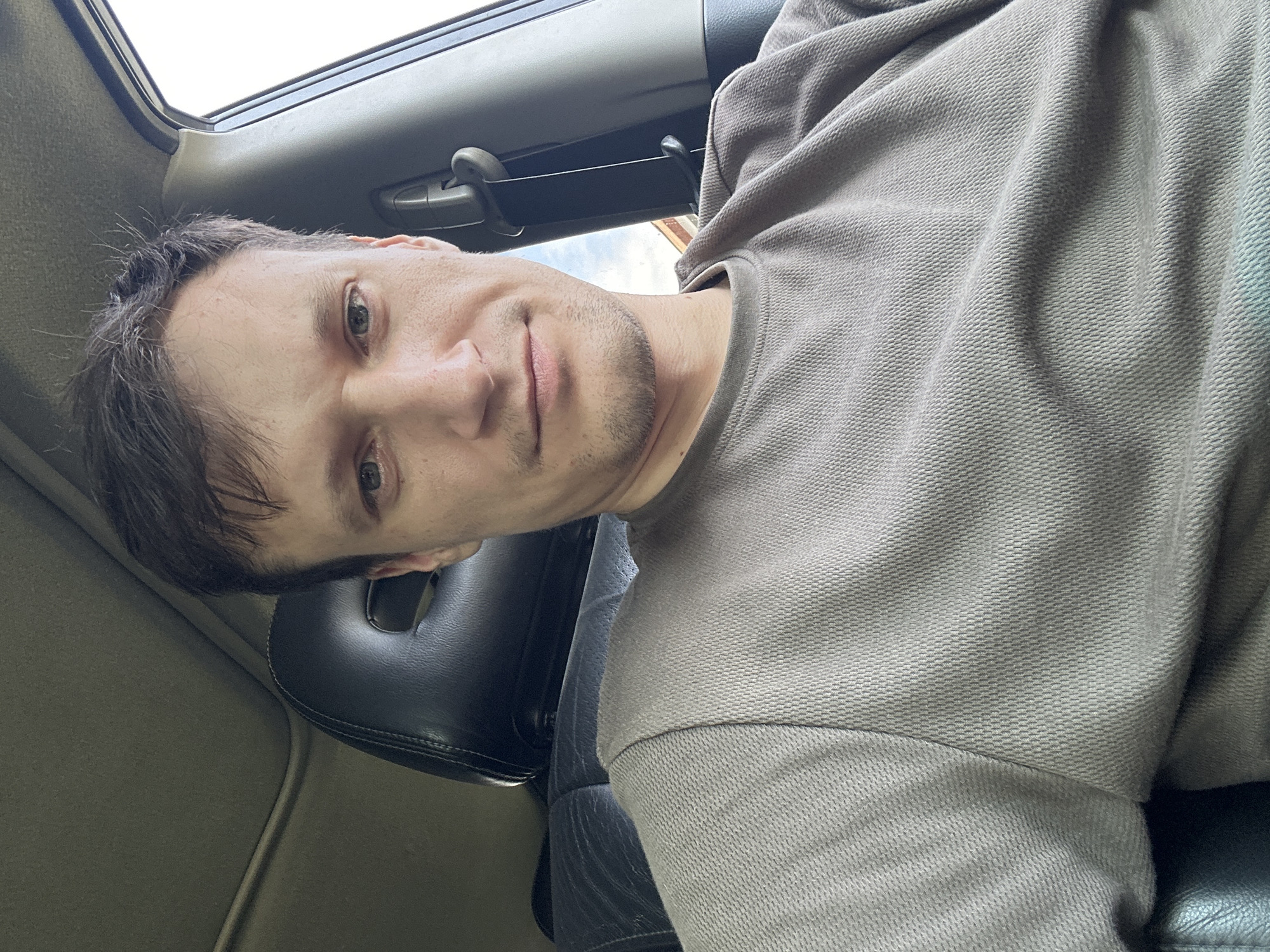 A close-up, selfie-style shot shows a fair-skinned man with short, dark brown hair and a light stubble on his chin. He is seated in the driver's seat of a car, with the interior of the vehicle visible, including the headrest of the passenger seat, the dashboard, and the roof of the car. The man is looking directly at the camera with a neutral expression. The background outside the car window suggests an overcast day. The scene is indoors, within a vehicle, in an unspecified urban or suburban setting.
