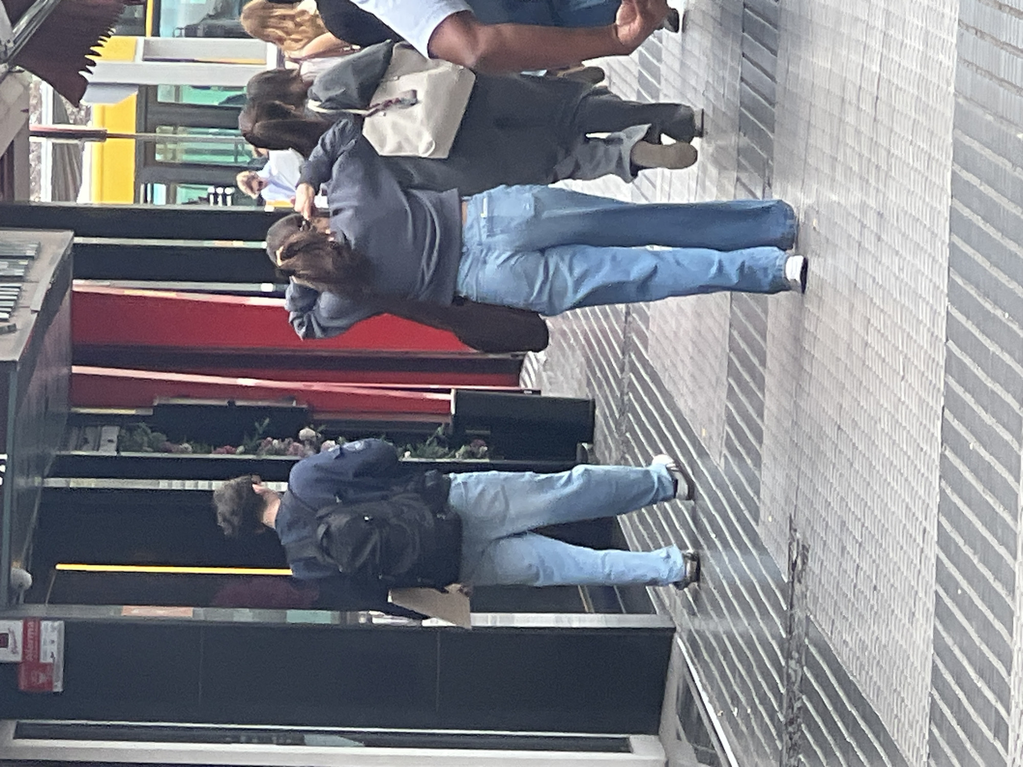 This image, likely captured in Las Palmas, Spain, depicts an outdoor urban scene bustling with pedestrians. The perspective is slightly elevated, looking down a paved street or walkway.
In the mid-ground, central to the frame, a person with dark, medium-length hair is seen from behind, walking away from the camera. They are wearing a dark blue long-sleeved hoodie or sweatshirt and light blue jeans. Their hands are raised near their head, as if adjusting their hair or reaching for something.
Further into the mid-ground, to the left of the central figure, another person is also walking away. This individual wears a dark top, light blue jeans, and white sneakers with dark accents. They have a black backpack on and appear to be holding a light-colored, possibly rectangular object in their left hand.
In the background, several other individuals are visible. One person wears a light grey top and darker pants, carrying a light-colored shoulder bag. Another, further back, has long, reddish-brown wavy hair and a dark top. More people are partially seen, indicating a busy public area.
The ground is paved with rectangular tiles, featuring distinct striped patterns of light and dark grey. To the left side of the frame, there's a dark-colored building facade, possibly a shop, with shelves visible through a window displaying greenery or decorative items. Above this, a red awning or canopy is present. Further down the street, a bright yellow structure, potentially part of a building or a vehicle like a bus, is glimpsed through reflections.
A small red sign with white text is affixed to the dark building facade; the word "ALERMA" is partially visible. The scene appears to be taking place during the daytime, with ample ambient light suggesting either an overcast day or indirect sunlight. No strong shadows are present. The overall atmosphere is that of a typical urban day, with people engaged in walking and going about their daily activities.