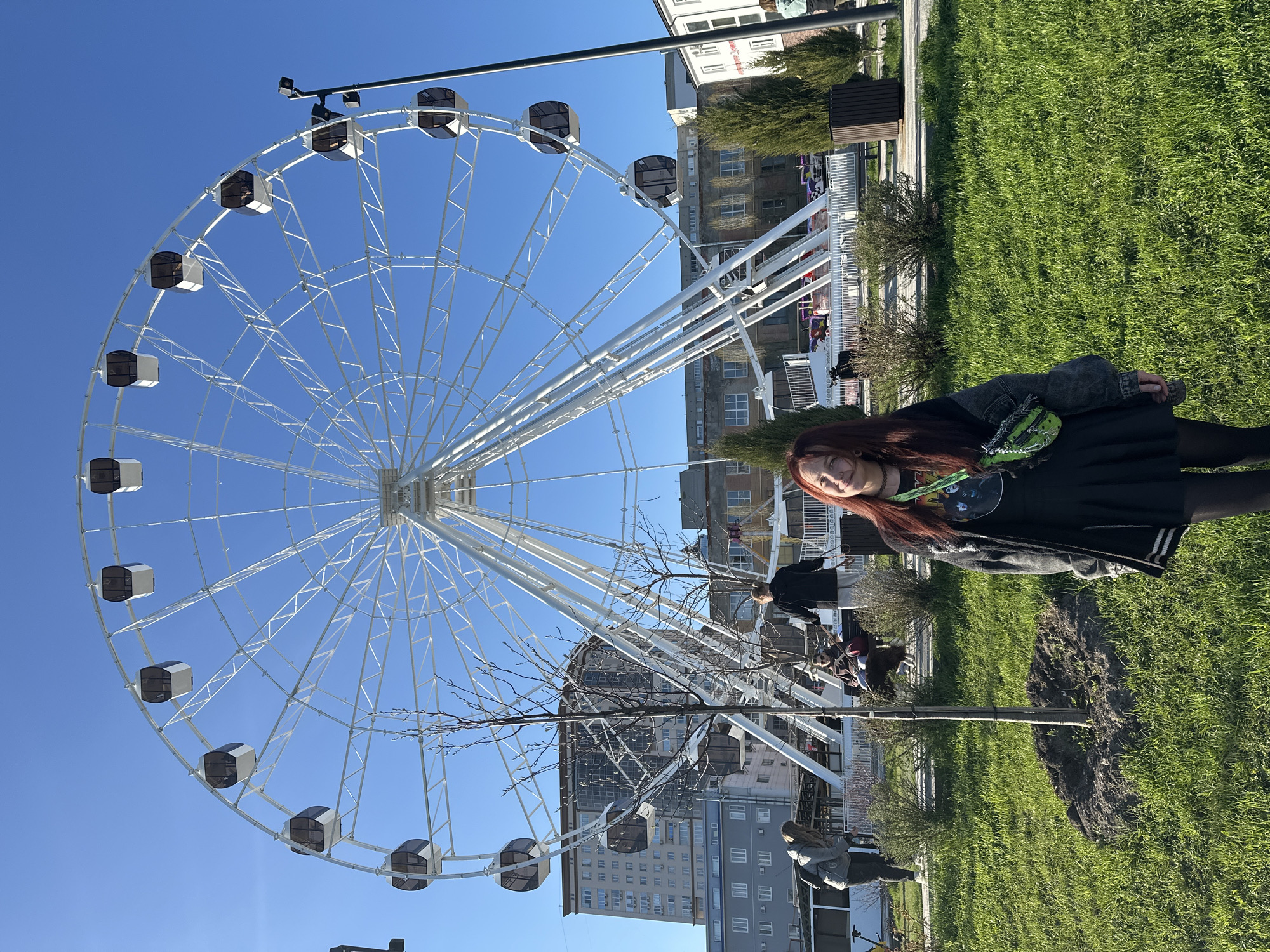 This media file captures a sunny outdoor scene in what appears to be an urban park or amusement area, located in Saratov, Russia.
**Subjects:** The central subject is a young woman with long reddish-brown hair, positioned on the right side of the frame. She is wearing a black jacket, a black skirt or dress, and black tights, accessorized with a bright green fanny pack or bag across her chest. She is looking towards the viewer with a slight smile. Dominating the left and central background is a large, white Ferris wheel featuring numerous dark-paned gondolas. Several other individuals are visible in the background, walking or sitting near the Ferris wheel. The scene also includes green grass, small trees (some with bare branches), and bushes. In the distance, various multi-story urban buildings form the backdrop.
**Activity or event taking place:** The main subject appears to be posing for a photograph. The overall setting, with the Ferris wheel and park environment, suggests a leisurely outing or visit to an amusement attraction. Other people in the background are engaged in typical park activities like walking or relaxing.
**Notable details:** The image was taken on a clear day with a bright blue, cloudless sky, indicating good weather conditions and ample sunshine. A black street light pole is visible at the top. On a distant white building, some blurry red text can be faintly discerned, though not clearly legible. The scene depicts an active public space. The image itself is presented in a landscape orientation, but the content suggests it was originally captured in portrait and rotated 90 degrees clockwise.