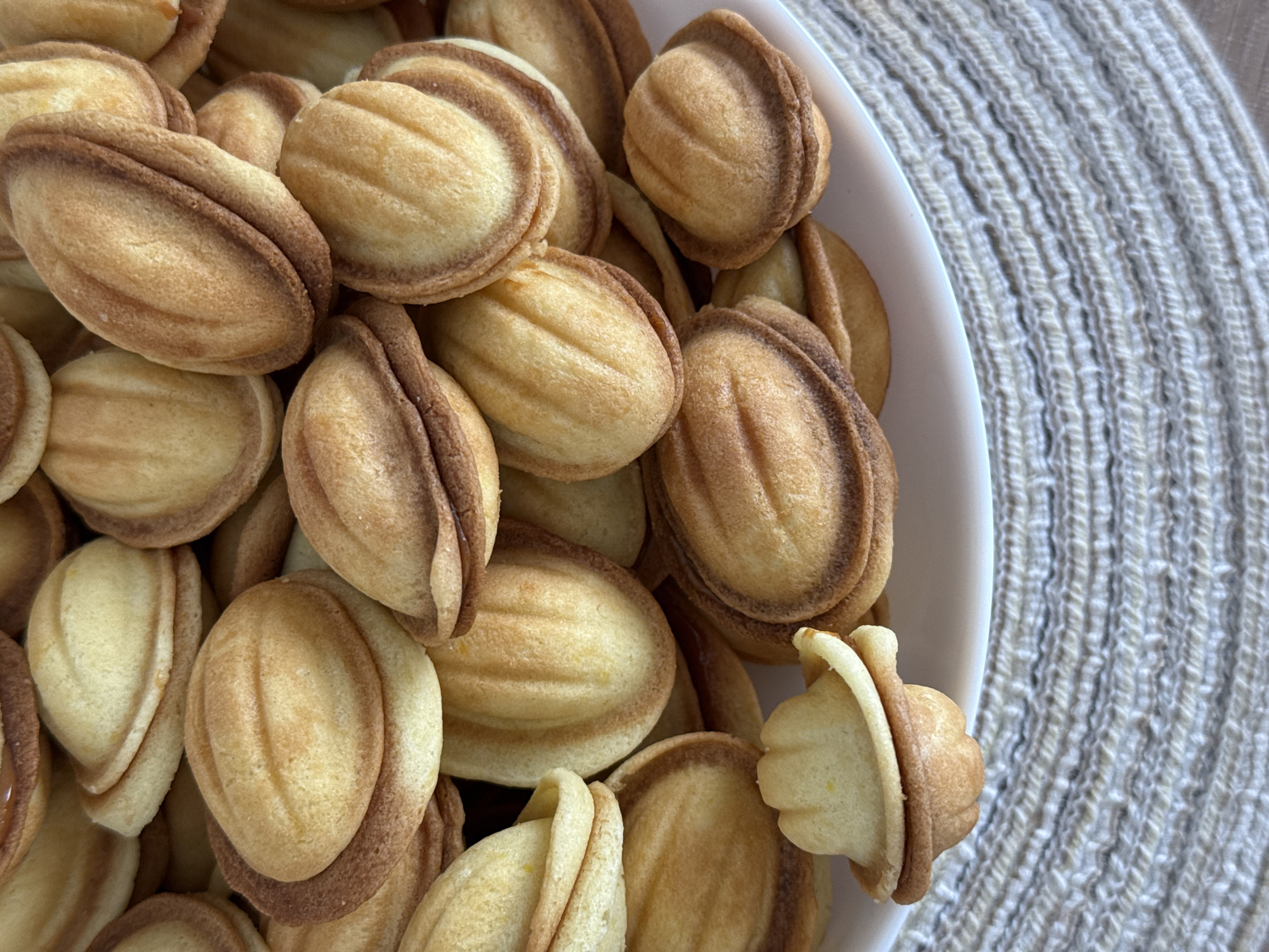 The media displays a close-up, high-angle view of a generous quantity of small, golden-brown cookies, likely a type of "nut cookie" or "Oreshki." These pastries are distinctly shaped, resembling walnut shells, with each cookie formed from two halves joined together. The predominant color is a light, toasted golden-brown, with some edges exhibiting a darker, more caramelized hue where the halves meet, enhancing their sculptural quality. Many cookies feature subtle longitudinal ridges or striations across their surfaces, indicative of the molding process.
The cookies are piled high within a white, ceramic bowl or dish, which is partially visible along the right edge and bottom of the frame, suggesting a large serving. The arrangement of the cookies fills the left and central portions of the image. To the right and upper right, a textured, woven mat or placemat serves as the background. This mat is light-colored, possibly off-white or light gray, and features a clear, regular pattern of raised, parallel lines from its coarse weaving.
The scene suggests an indoor setting, possibly a kitchen or dining area, with soft, ambient lighting. There are no people present, and no specific time of day or location is discernible. The cookies appear ready for serving or consumption. No visible text is present in the image.