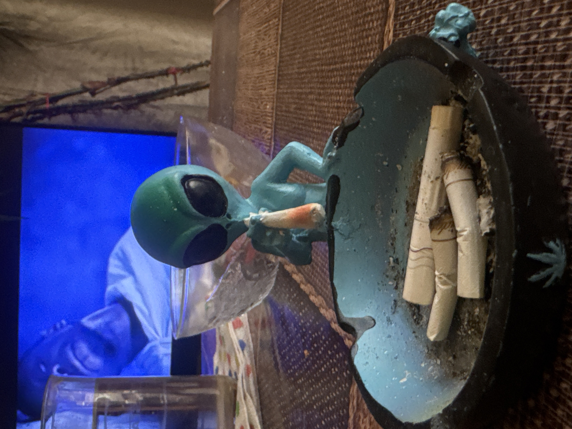The image shows a close-up, eye-level shot of an alien figurine with a cigarette in its mouth, positioned in an ashtray filled with several cigarette butts. The figurine is turquoise and appears to be made of plastic or resin. It has large black eyes and a slender body. The ashtray is dark blue with a lighter blue rim and a decorative green leaf design at the bottom. Behind the ashtray and figurine, a glass bowl is visible, partially filled with what appears to be water or ice. To the left, a glass filled with a clear liquid sits on a patterned cloth. In the background, a television screen displays a blue hue with a faint image of a person's face. The scene is set on a woven, brown surface, possibly a table. The overall environment suggests an indoor setting. There is no information in the image to suggest a location in Sofia, Bulgaria.