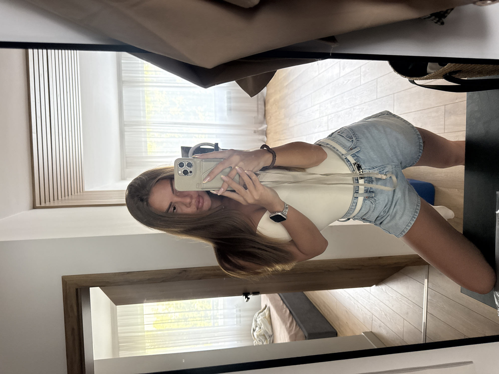 A young woman with long, brown hair takes a mirror selfie in a bedroom. She is wearing a white bodysuit and light blue denim shorts with a white belt. She is holding a smartphone in her right hand, with her left hand resting on her hip. Her left leg is bent at the knee, and she is leaning forward.
The room is modern and well-lit, with light wood flooring and white walls. A large mirror dominates the right side of the frame, reflecting the interior of the room. In the reflection, a doorway leads to another room with a bed and sheer curtains covering a window. A brown leather jacket is draped over a stand to the right of the mirror. The overall impression is of a casual, stylish personal moment captured within a contemporary living space. It is not possible to determine the specific location within Plovdiv, Bulgaria from the visual information provided.
