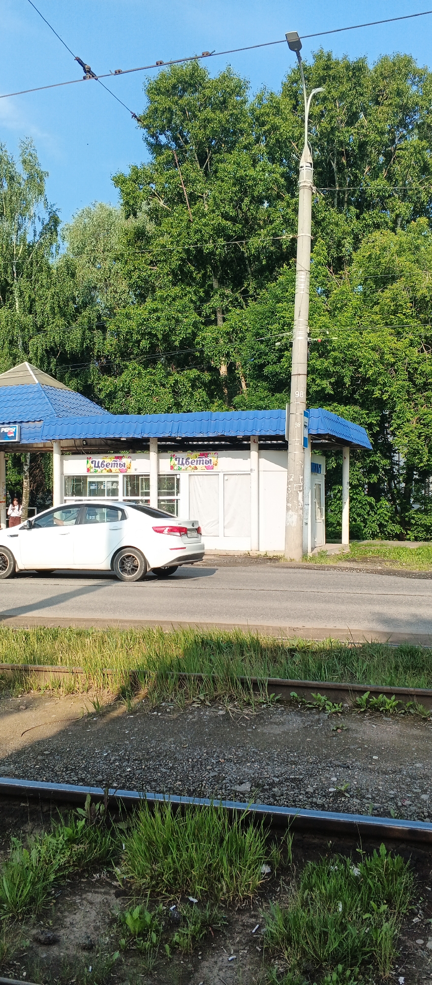 The image captures a view of a street in Izhevsk, Russia, featuring a small pavilion with a blue roof, likely a flower shop, indicated by signs reading "Цветы" (Flowers). A white car is driving past the pavilion on the asphalt road. In the foreground, tram tracks are visible, bordered by overgrown grass and gravel. A concrete lamppost stands to the right of the pavilion. Dense green trees form a backdrop behind the structures and the road. The sky is clear and blue, suggesting a sunny day.