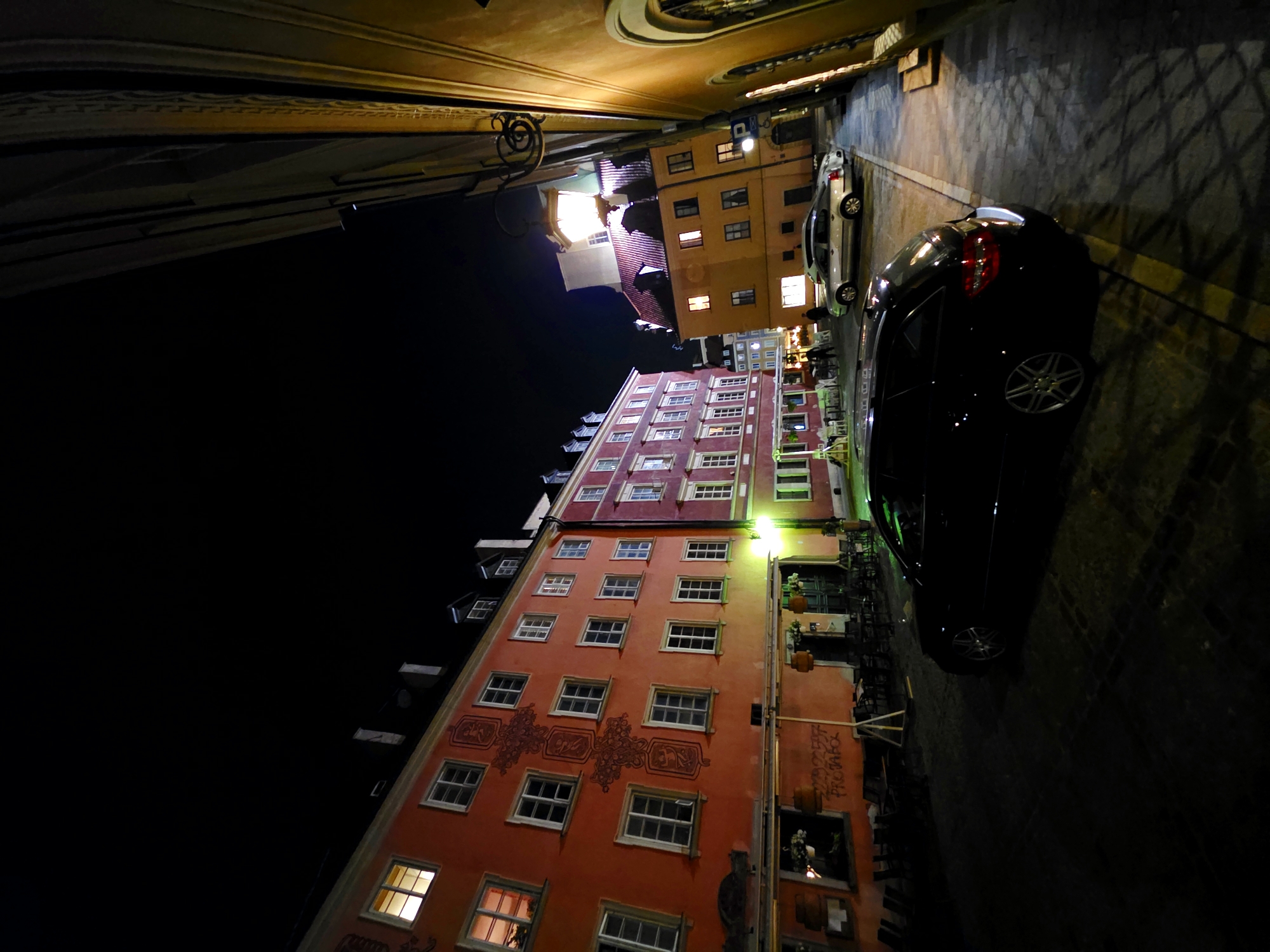 The image depicts a nighttime street scene in Warsaw, Poland, likely the Old Town, characterized by historic architecture and cobblestone pavement. A black station wagon is parked prominently in the foreground, its rear illuminated by streetlights. Beyond the station wagon, several buildings with illuminated windows line a narrow street. The building on the left is painted a vibrant orange with decorative red motifs on its facade. Across the street, a yellow building is visible, with a large, ornate lantern hanging from an external bracket. Several other cars are parked further down the street. The scene suggests a quiet evening in a well-preserved historical district.