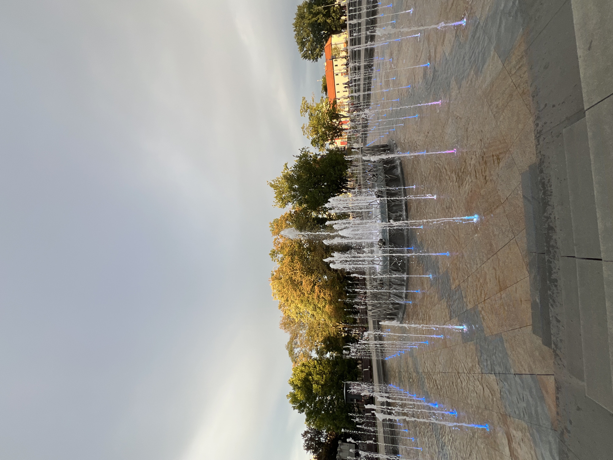 The image shows a wide-angle view of a modern fountain plaza in the city of Lublin, Poland. The fountain features numerous jets of water, many of which are illuminated with blue and pink lights. The water sprays create dynamic vertical lines against the backdrop of a cloudy sky. The plaza is paved with large, square tiles of varying colors, including shades of tan, brown, and gray, arranged in a patterned design. In the foreground, there are concrete steps leading up to the plaza. Behind the fountain, a line of trees with autumn foliage in vibrant shades of green, yellow, and orange is visible. Further in the background, a building with an orange tiled roof and white walls, possibly a historical structure, stands against the sky. Several indistinct figures of people can be seen in the distance near the building, and some park benches are also present among the trees. The overall scene captures a public space in Lublin, likely a park or a pedestrian zone, enhanced by a contemporary water feature and illuminated at dusk or during a gray day.