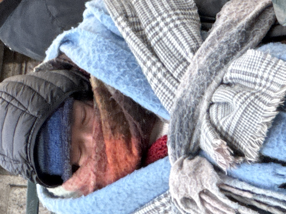 A person, covered in layers of blankets and scarves, is sleeping. The person wears a black puffy jacket and a blue beanie. An orange and pink patterned scarf is wrapped around their face. They are cocooned in a light blue blanket and a grey patterned scarf with tassels. The background is blurred.