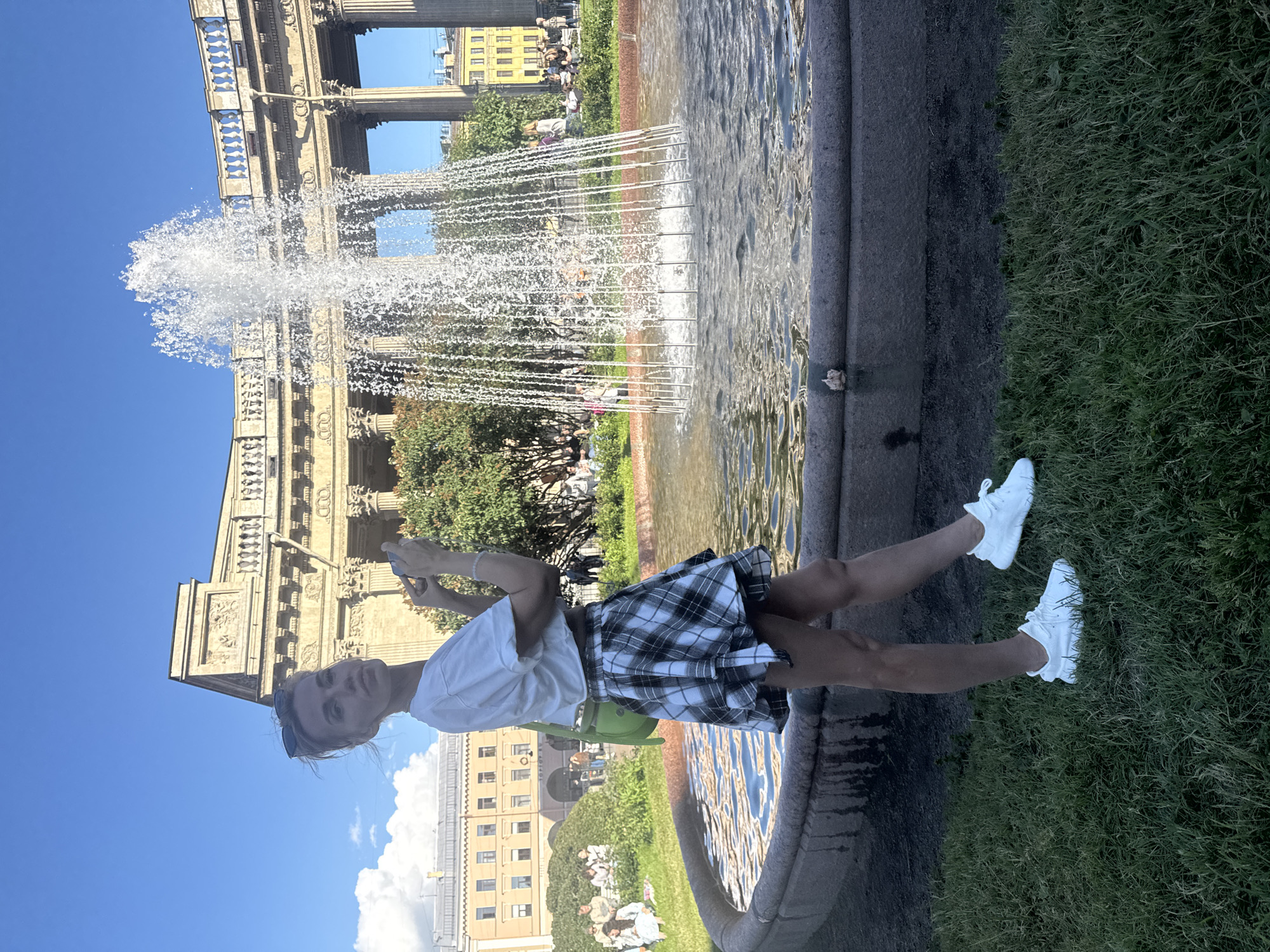 A young woman in a white t-shirt and a plaid skirt poses in front of a fountain. She is holding a smartphone in her right hand. The fountain sprays water high into the air, with a large building in the background. The building appears to be made of stone and has classical architectural elements, including columns and ornate carvings. The sky is clear and blue, suggesting it is a sunny day. The scene is set in a park-like area with lush green grass. In the background, other people can be seen relaxing on the grass. The overall atmosphere seems peaceful and relaxed, with the woman appearing happy and engaged with her phone. The location is identified as Saint Petersburg, Russia. The time of day appears to be daytime.