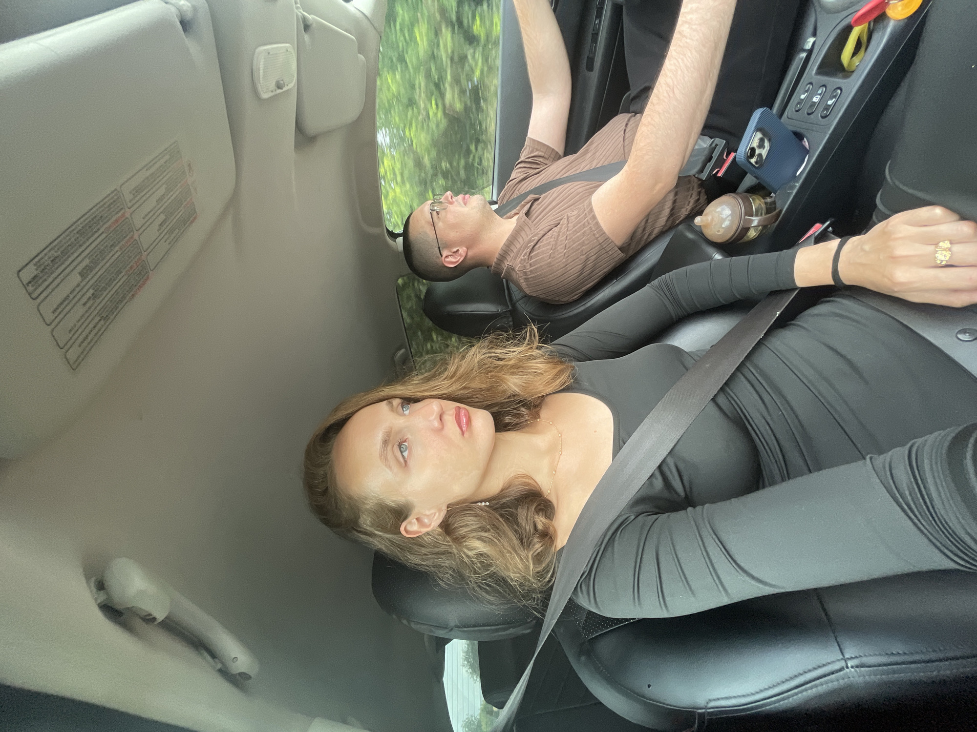 The image shows two individuals in the front seats of a car. A woman with blonde hair is in the driver's seat, while a man with short dark hair and glasses is in the passenger seat, driving. Both are wearing seatbelts. The woman is wearing a black long-sleeved dress and a necklace. The man is wearing a brown short-sleeved collared shirt. In the center console, a blue phone, a sippy cup, and other items are visible. The background outside the car shows blurred green trees, suggesting the vehicle is in motion. The scene appears to be a road trip or a commute. The location is specified as Pirogovo, Russia.
