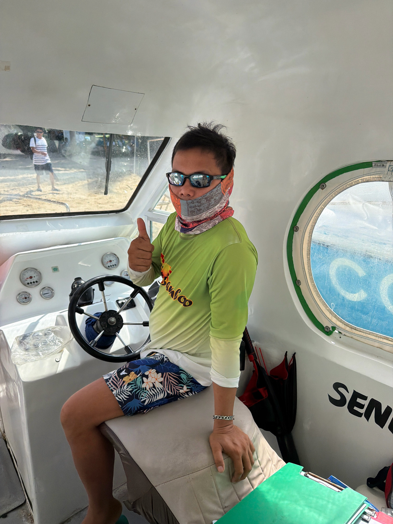 A man sits in the captain's seat of a small white boat, holding up a thumbs-up gesture. He is wearing a green long-sleeved shirt with a colorful bandana covering his lower face and sunglasses. His shorts feature a tropical floral print. In the background, through a large window, another person in a striped shirt and dark shorts stands on a sandy beach. The boat's interior shows a steering wheel, various gauges, and a circular porthole with a blue inscription visible through it. The boat is docked, or moving slowly, near the shore of a beach in Nabas, Philippines.