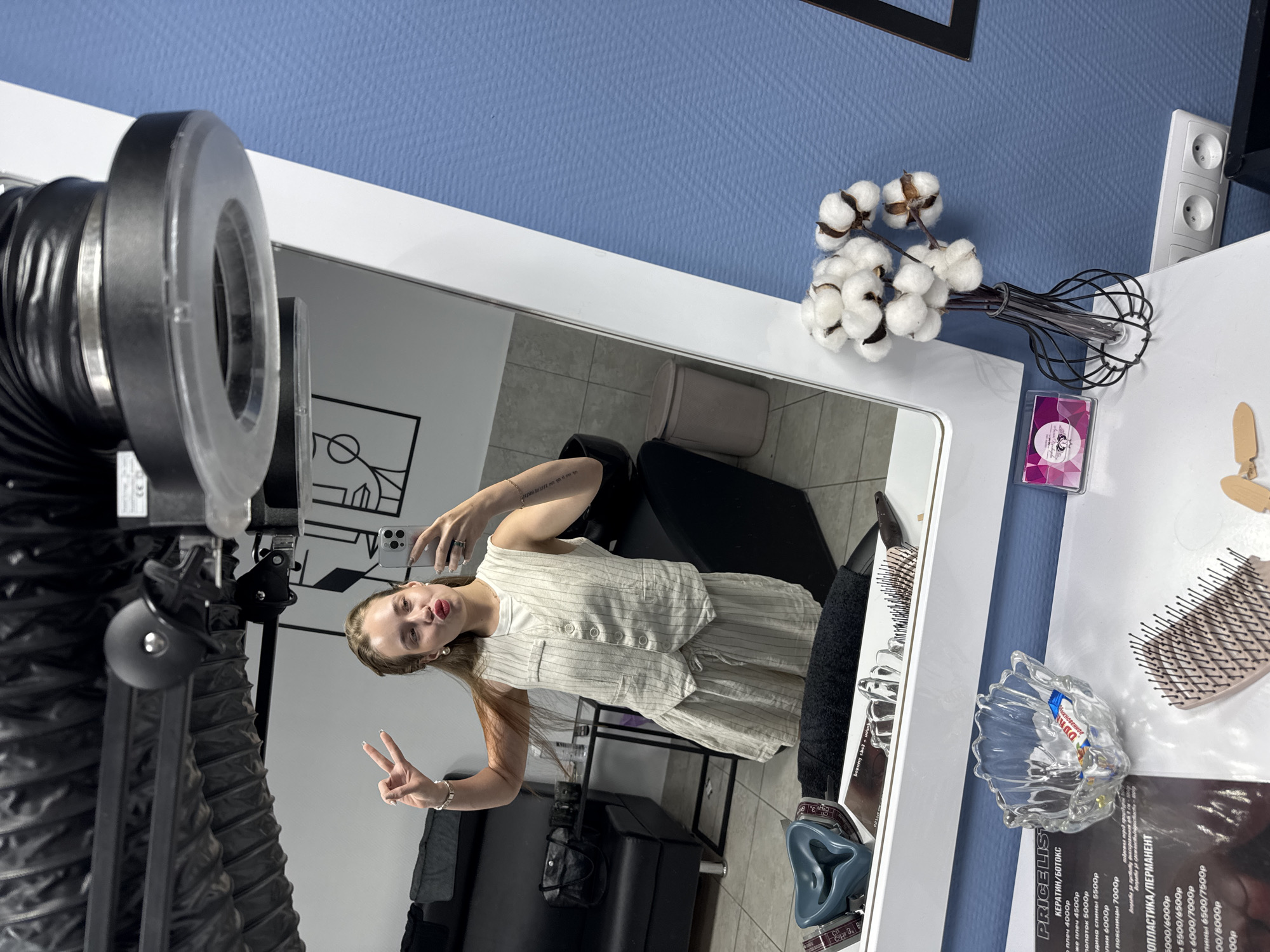 A young woman is taking a selfie in a salon, likely in the city of Chaikovsky, Russia, based on the Cyrillic text in the price list visible in the reflection. She is standing in front of a large, rectangular mirror, holding her phone up. She is wearing a light-colored, striped vest and shorts set, with a white top underneath. She is holding up a peace sign with her left hand and has a playful expression on her face.
The reflection in the mirror shows her, the salon interior, including a black chair, and a wall with a graphic design. To the left of the mirror is a professional lighting rig, and to the right is a white surface with various items including a bouquet of cotton flowers in a black wire vase, a hairbrush, a clear glass dish with what appears to be sugar packets, and a price list with text in Russian. The wall behind the mirror is a light blue color. The overall environment suggests a modern and well-lit salon space.