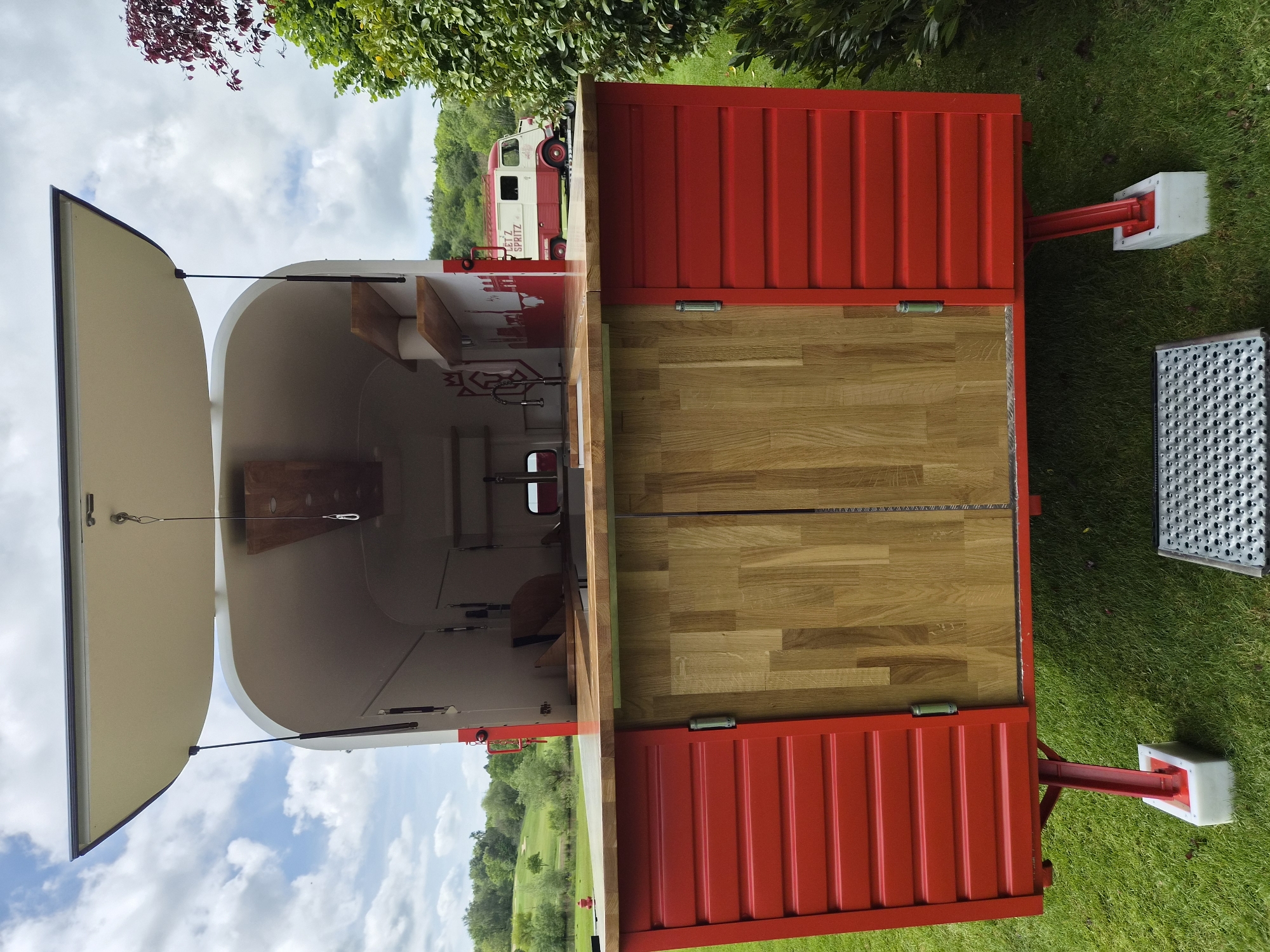 The image shows a red and white mobile bar, likely a converted shipping container, parked on a grassy area. The bar has a wooden countertop, open on one side to reveal the interior. The interior appears to be a food or beverage preparation area with shelves and equipment. A vintage red and white van is partially visible in the background, suggesting a mobile catering or beverage service. The location is on grass in Lenningen, Luxembourg.