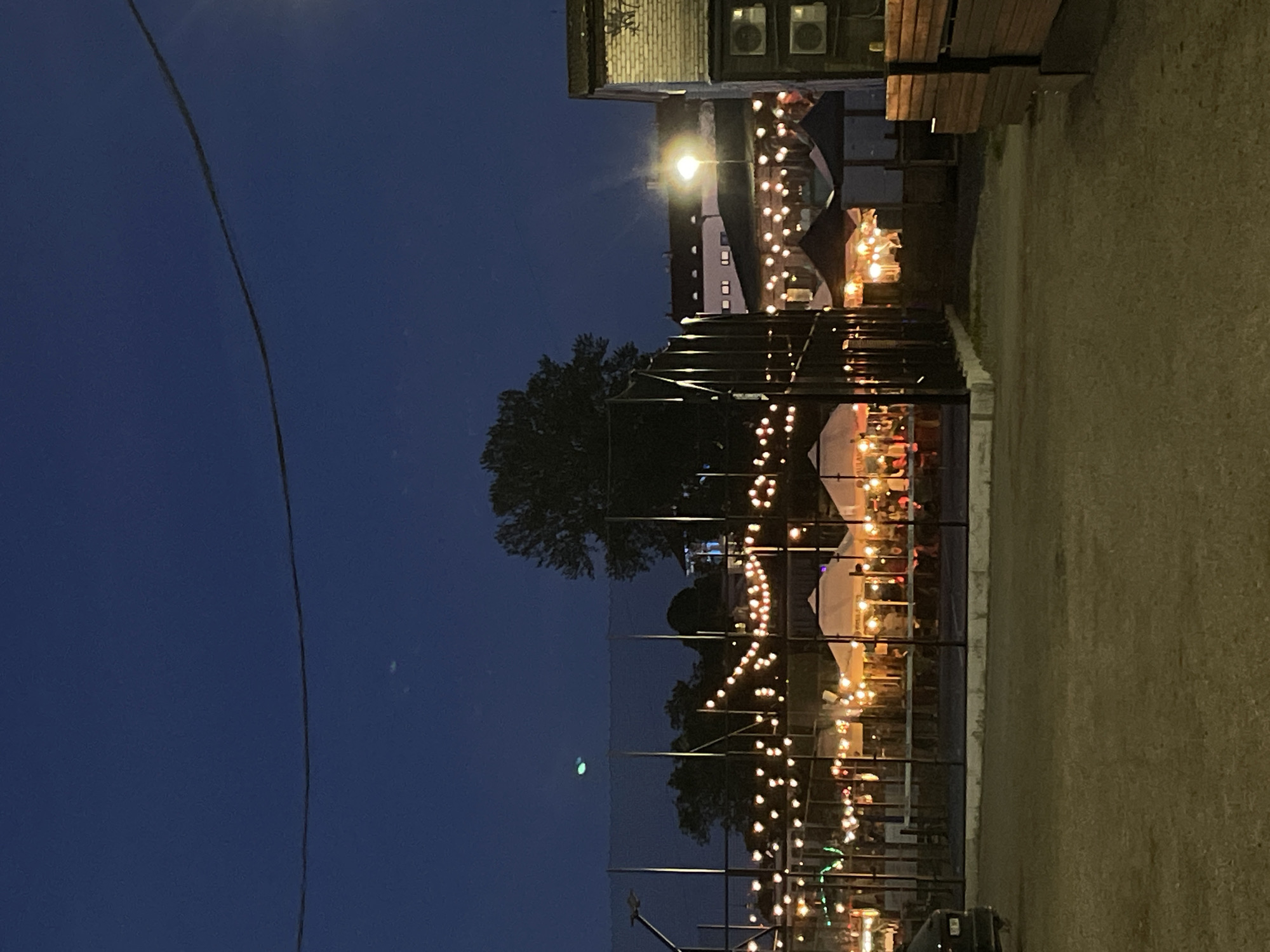 The image depicts an outdoor scene at dusk or night, likely in Tallinn, Estonia, given the presence of strung lights and what appear to be market stalls or an outdoor dining area. The sky is a deep blue, transitioning into darkness. In the foreground, there is a gravelly or dirt-covered area, and to the left, a portion of a dark-colored vehicle is visible.
Dominating the mid-ground is a fenced area, possibly a sports court or an enclosed event space, lined with numerous small, warm-toned lights strung overhead. Inside this fenced area, several white and dark-colored tents or marquees are set up, illuminated from within and by the surrounding string lights. Tables and chairs are visible beneath these structures, suggesting an area for dining or gathering. People are present within these tents, but their features are not discernible due to the distance and lighting.
In the background, buildings with illuminated windows are visible, along with streetlights. A large tree with a full canopy stands prominently behind the fenced area. To the right of the frame, a brick building with external air conditioning units is present, adjacent to a wooden structure. A single bright streetlight illuminates the scene from a middle-ground building. The overall impression is of a lively, illuminated outdoor venue operating after dark.