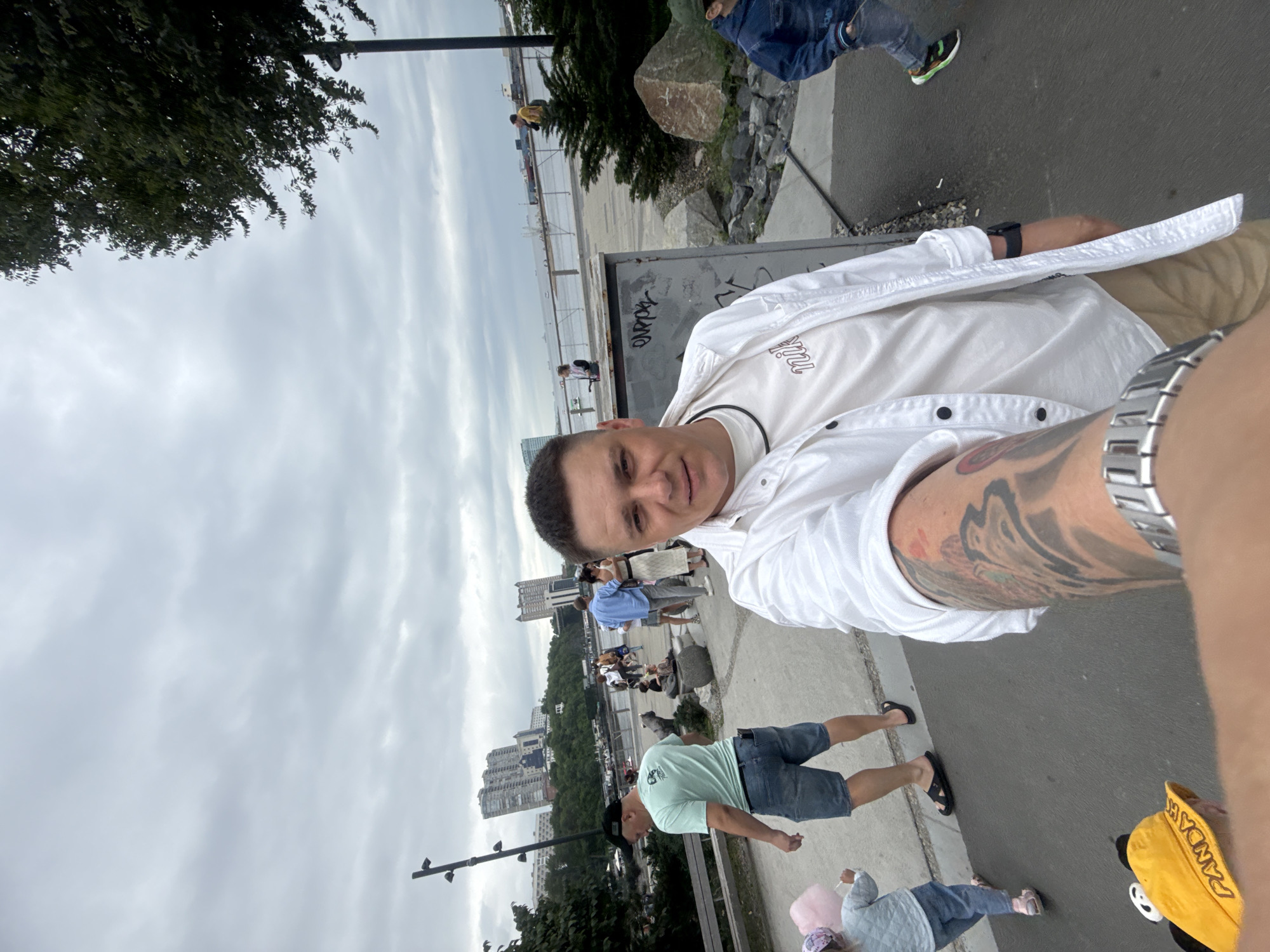 A man in a white shirt and beige shorts takes a selfie outdoors. He has tattoos on his arm and is wearing a watch. In the background, people are walking, and a child is holding cotton candy. The scene appears to be in a park or promenade with buildings and a body of water visible in the distance. The sky is cloudy. This location is consistent with the waterfront area of Vladivostok, Russia, possibly near the Golden Horn Bay.