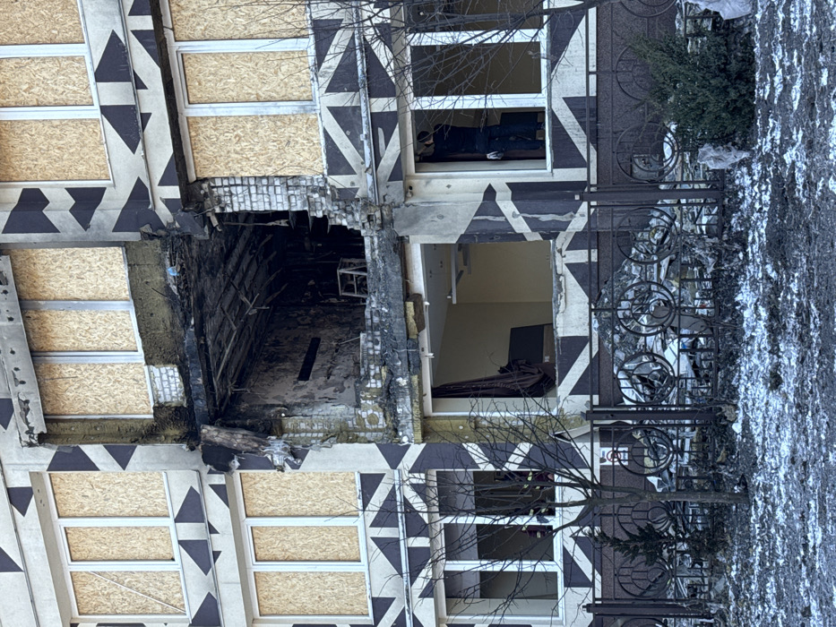 The image displays a multi-story building in Kyiv, Ukraine, exhibiting significant damage. The building's facade is grey with a distinctive black geometric pattern of triangles and diagonal lines.
A large, irregular opening marks the central section of the building, spanning at least two floors. The upper portion of this opening reveals a deeply charred and blackened interior room, consistent with fire damage. Structural elements are visible within this cavity, and a light-colored, possibly wooden, stool or small table is present amidst the debris inside. The ceiling above this upper damaged room appears compromised or collapsed. The lower portion of the central damage opens into a less damaged interior room with light-colored walls, an intact ceiling, and a dark curtain partially visible.
Numerous windows across the building's facade, particularly on the upper levels and some on the lower, are boarded up with light-colored plywood or particle board. Exposed brickwork and concrete are visible around the damaged sections.
In a lower-level window on the right side of the frame, one individual, dressed in dark clothing, is seen standing inside, appearing to be working near the window frame. Through a lower-left window, several figures in what appear to be red jackets are faintly discernible.
The foreground features a black metal fence with circular decorative elements. Behind this fence, directly below the damaged building sections, lies a substantial pile of debris, including shattered glass and various building materials. The ground in the foreground is a mixture of dark soil, scattered debris, and patches of melting snow. Several bare deciduous trees and small evergreen shrubs are also present near the fence line.