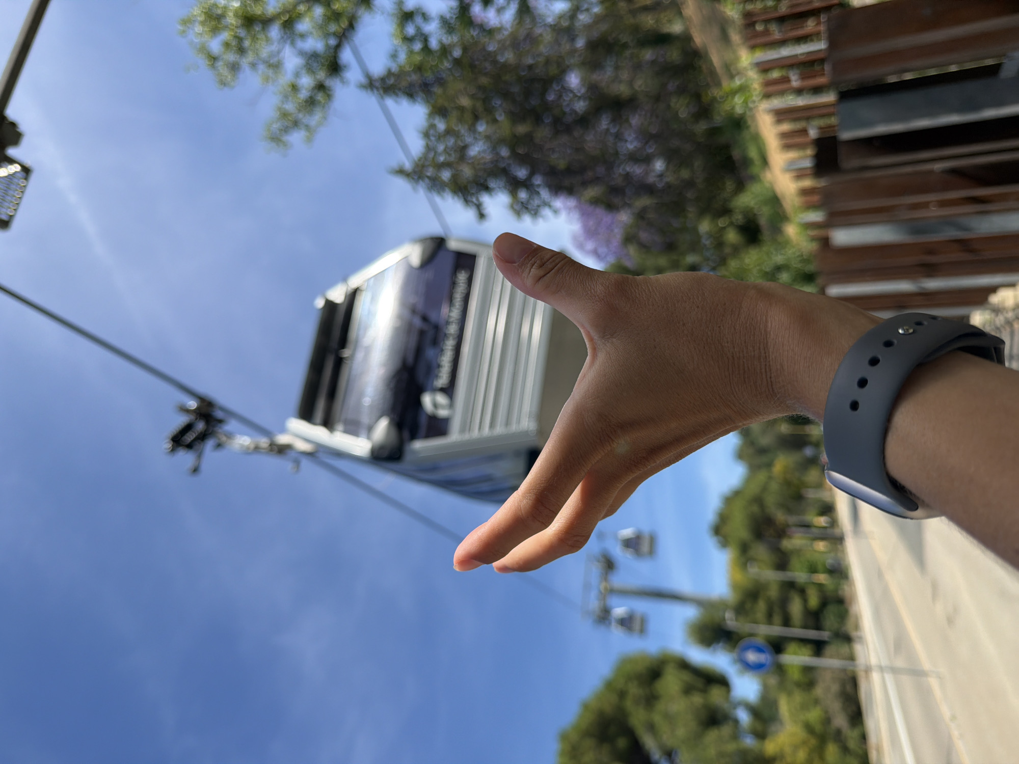 A first-person perspective shot shows a hand with a smartwatch on the wrist reaching towards a cable car in the city of Barcelona, Spain. The hand is in focus, with fingers slightly spread. The cable car, labeled "TRANSBASCARANS", is suspended from a cable and is also in focus. In the background, more cable cars are visible along the same line, as well as trees and a bright blue sky. A road with lampposts and a traffic sign can be seen below. A wooden fence runs along the right side of the frame.