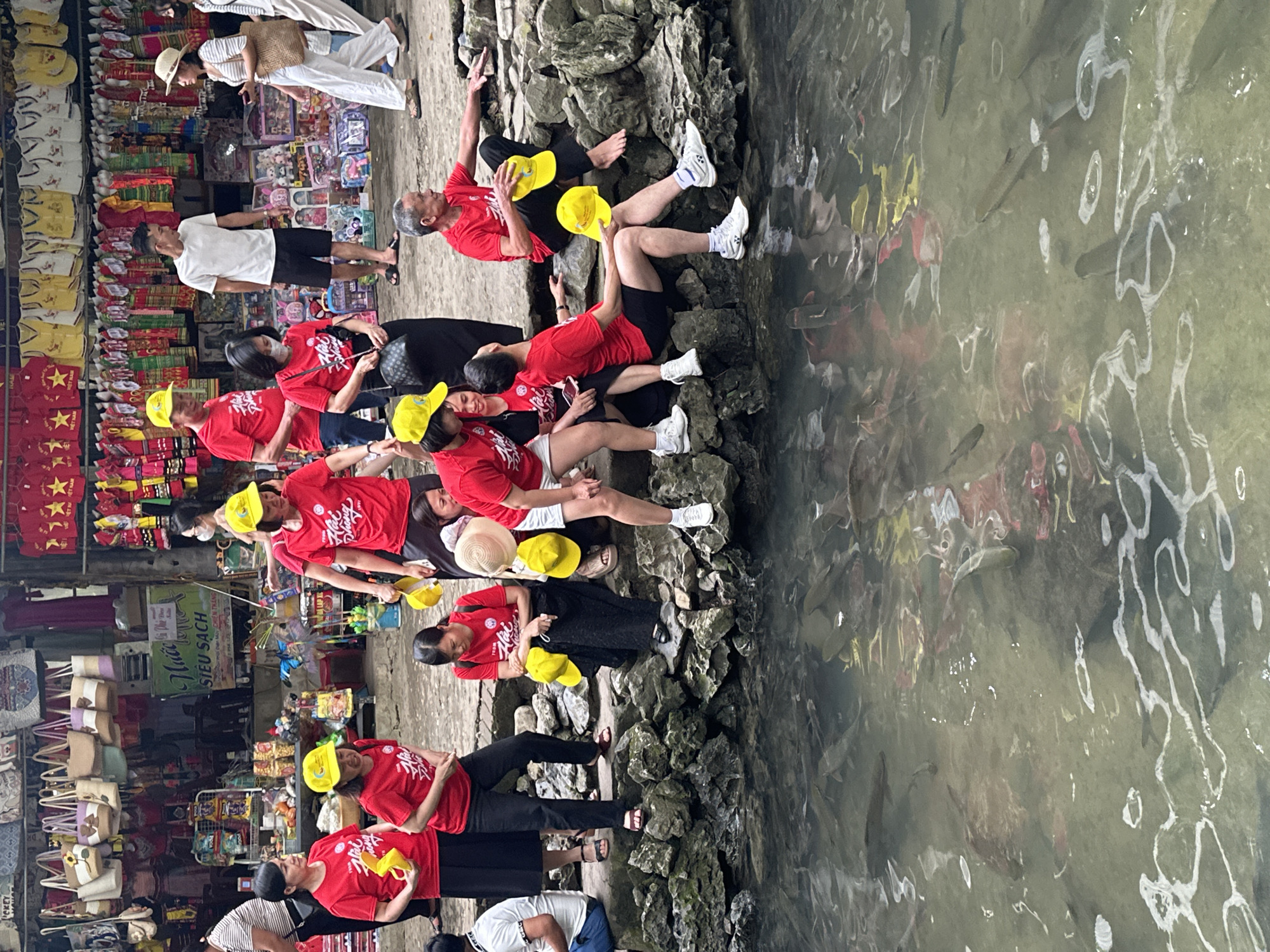 In Cẩm Tú Commune, Vietnam, a group of people, predominantly wearing red shirts and yellow hats, are gathered by a waterway. The individuals are positioned on a rocky embankment and in the shallow water, observing and interacting with numerous fish swimming near the surface. Some are seated on the rocks, while others are standing or crouched closer to the water's edge. The background reveals a marketplace with stalls displaying clothing and various merchandise. The atmosphere appears to be casual and observational, with a focus on the aquatic life.