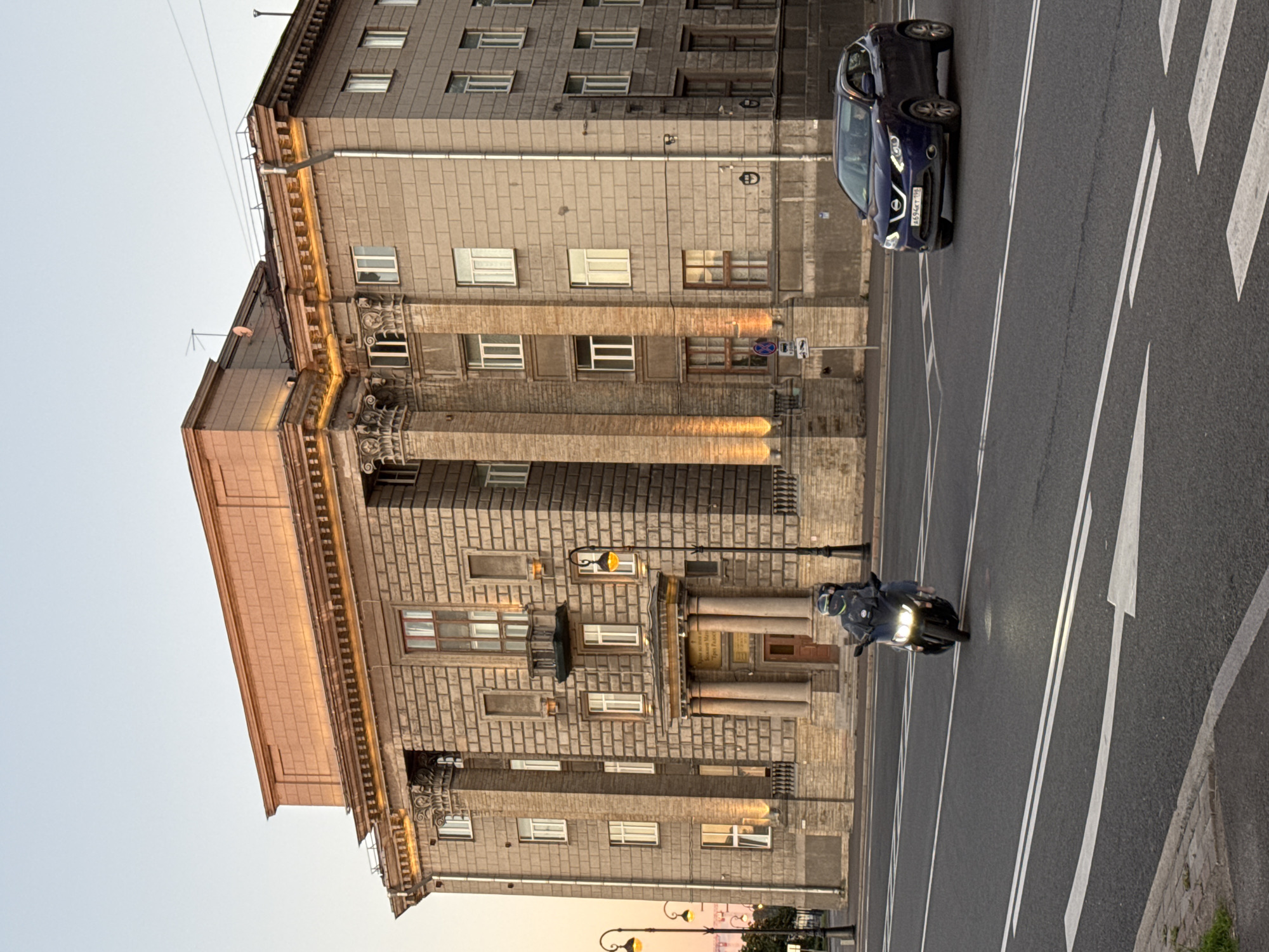 The image shows a multi-story building with classical architectural elements, located in Saint Petersburg, Russia. The building features large columns, decorative cornices, and numerous windows. The facade is constructed from stone blocks, and the upper portion is illuminated by warm lighting. In the foreground, a dark-colored car is driving on a multi-lane road. A motorcyclist wearing a helmet and riding a black scooter is also on the road, heading towards the camera. The sky is clear with a soft, pale hue, indicating it might be early morning or late evening. Streetlights are visible on the sidewalk next to the building.