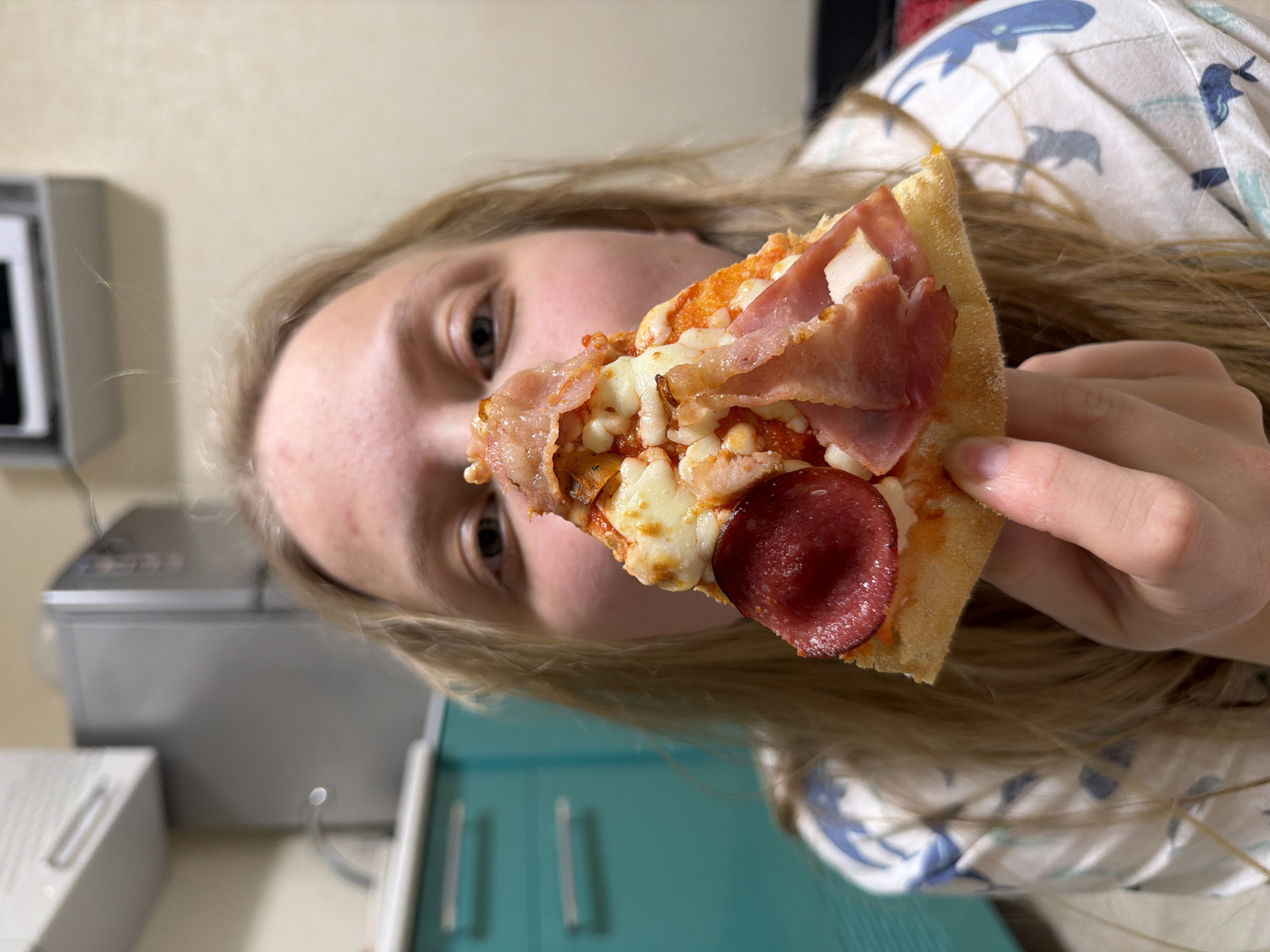 A blonde-haired person holds a slice of pepperoni pizza in front of their face. The person is wearing a white shirt with blue whale patterns. The background includes a gray kitchen appliance and a blue cabinet. There is no evidence to suggest the location is Izhevsk, Russia.