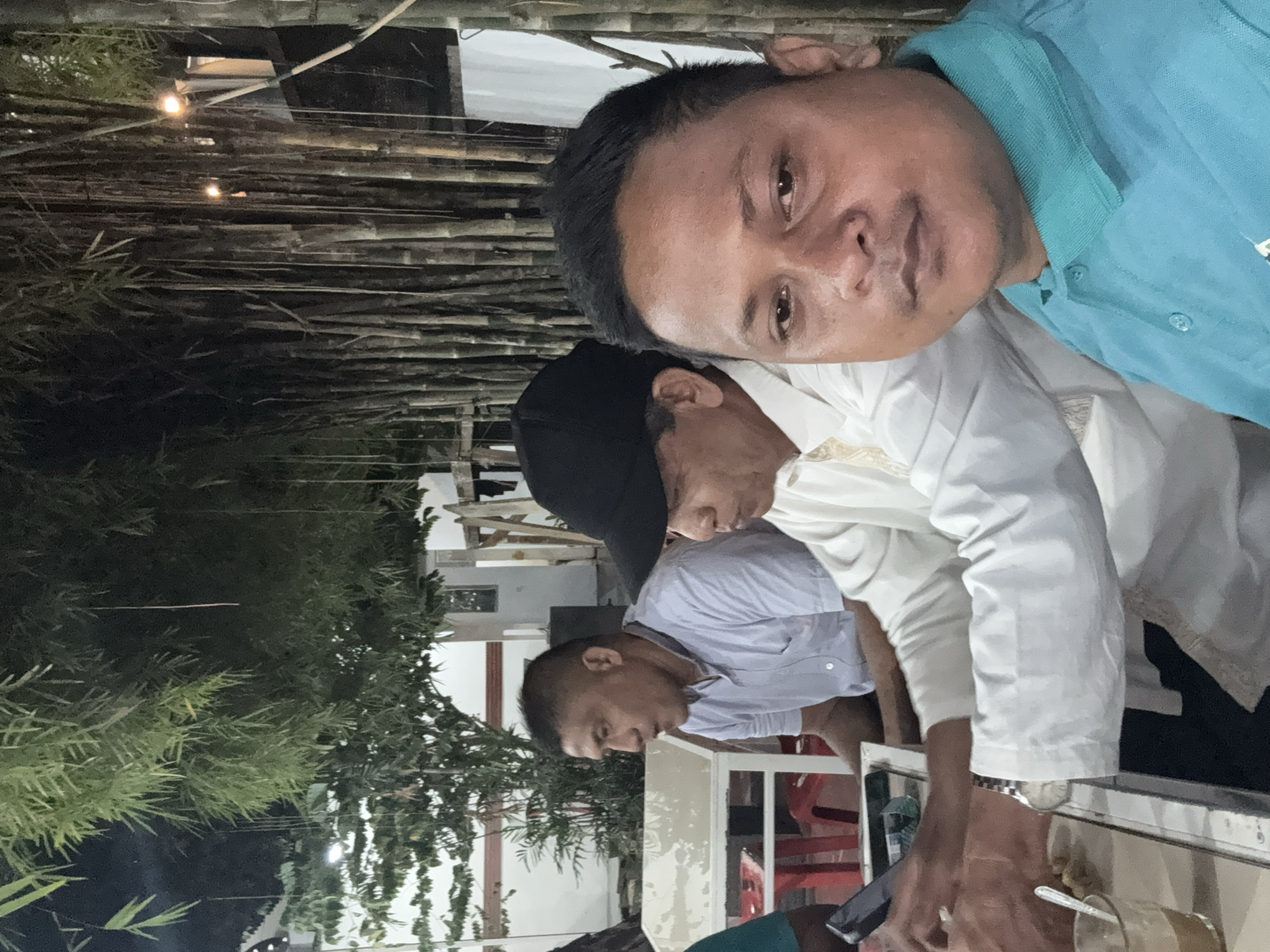The image captures an informal gathering in Lhokseumawe, Indonesia. Three men are visible, all wearing casual clothing. One man is wearing a light blue shirt, the second wears a white shirt, and the third is wearing a light blue button-up shirt and a black cap. The men are likely sitting at an outdoor dining area, as evidenced by the table and chairs in the foreground. A glass of liquid sits on the table. Background elements show a residential structure, trees, and what appears to be some sort of bamboo-themed wall or roof. The scene is lit by what seems like artificial light.