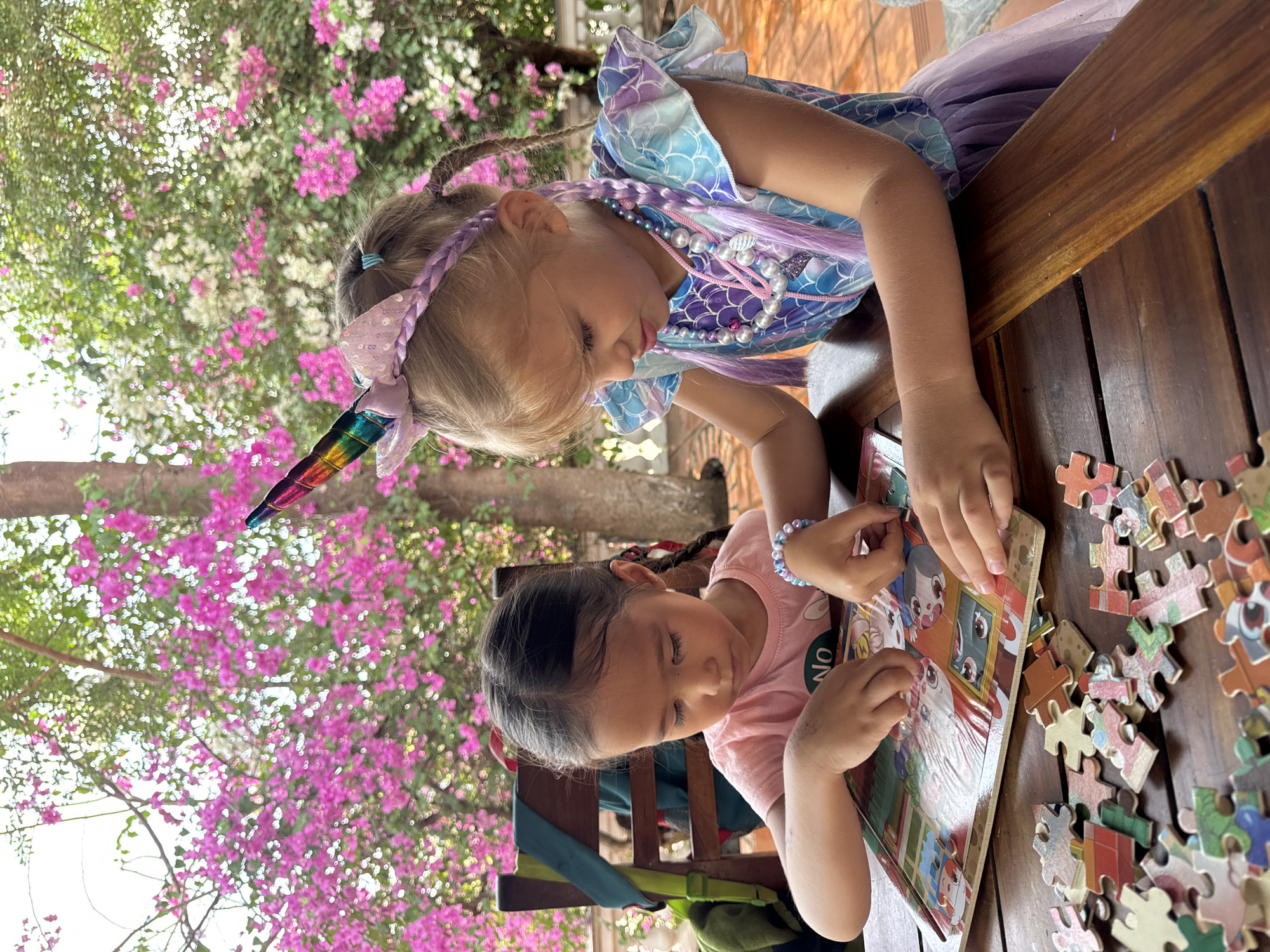 Two young girls are sitting at a wooden table, working on a puzzle. One girl, with blonde hair braided and adorned with a unicorn headband, wears a blue and purple mermaid-themed dress with a pearl necklace. The other girl, with brown hair in braids, wears a pink t-shirt. They are both focused on the puzzle, which is laid out with its picture in front of them and loose pieces scattered on the table. The scene appears to be outdoors, possibly on a patio, with a backdrop of vibrant pink bougainvillea flowers. The image suggests a casual, sunny day in Mũi Né, Vietnam, where the shot was captured.