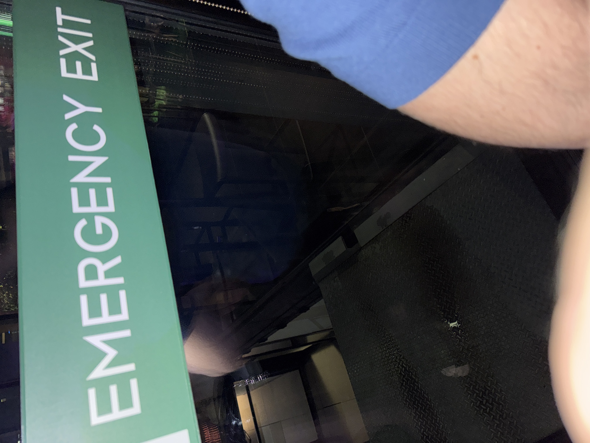 A close-up, oblique-angle shot shows a green "EMERGENCY EXIT" sign affixed to what appears to be a glass partition or window. The white lettering is vertically oriented on the sign. Through the glass, a dimly lit environment is visible, with indistinct shapes suggesting interior spaces. The arm and shoulder of a person wearing a blue shirt occupies the upper portion of the frame, obscuring some of the background. To the right of the glass partition is a dark, textured surface, possibly flooring or paneling. The location of the scene, city, and country are not discernible from the provided image.