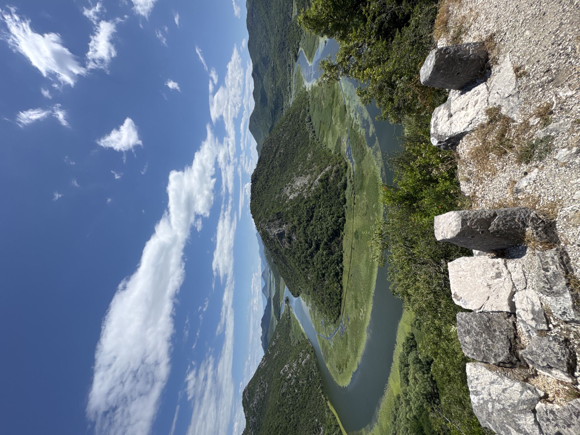 A scenic overview captures a winding river flowing through a lush, green valley. The river, a dark, reflective ribbon, is bordered by dense vegetation, with patches of lighter green aquatic plants scattered across its surface. The valley is flanked by rolling, tree-covered hills that recede into the distance. The sky above is a vibrant blue, dotted with fluffy white clouds. In the foreground, a stone wall, constructed from large, irregularly shaped blocks, sits atop a gravelly embankment. The wall appears weathered and aged, suggesting historical significance. The overall scene depicts a tranquil and natural landscape under a bright, sunny sky.