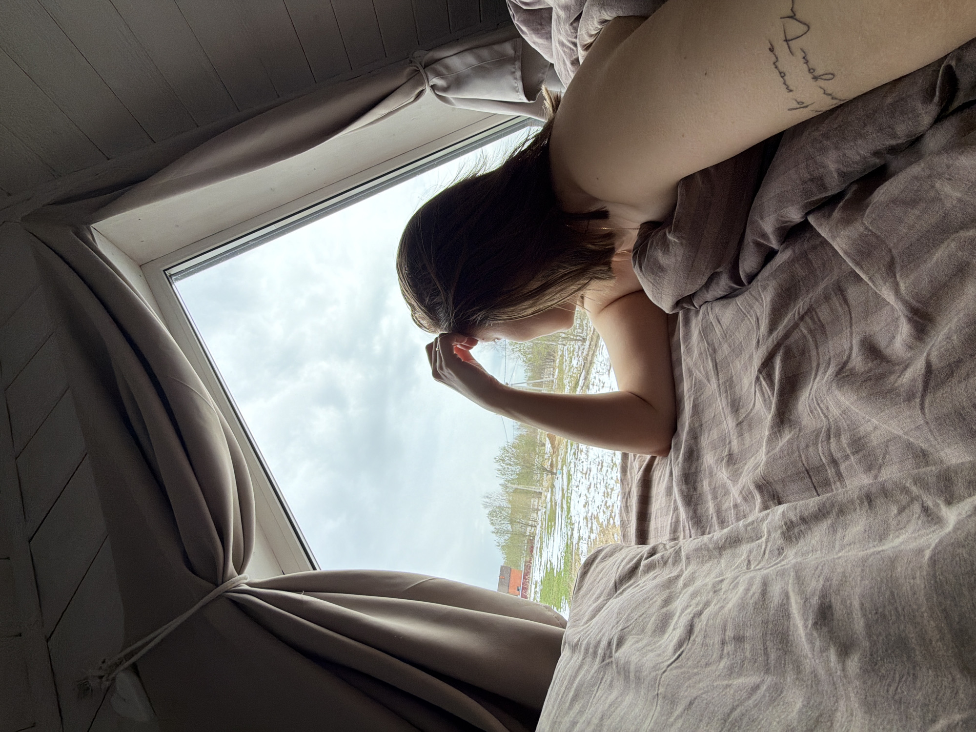 The image captures a person resting in bed, looking out a large window on an overcast day in what appears to be Бунятино, Russia. The perspective is from above and to the right, showing the person partially covered by light brown/grey bedding. Their head is turned towards the window, with their dark hair falling around their face as they gaze outside.
The window, possibly a skylight or positioned high, reveals a cloudy sky above a landscape with patches of snow and grass, bare trees, and a small, red-roofed building in the distance. The interior features light-colored, panelled walls or ceiling, and matching light brown/grey curtains tied back on the right side with a white cord.
A notable detail is a cursive tattoo on the person's left upper arm, which includes partially visible text, seemingly in French, such as "mon cœur" (my heart) and other phrases that are not fully decipherable. The scene suggests a quiet, contemplative moment, possibly during late winter or early spring, given the weather and bare trees. The time of day is daytime, indicated by the natural light filtering through the clouds.