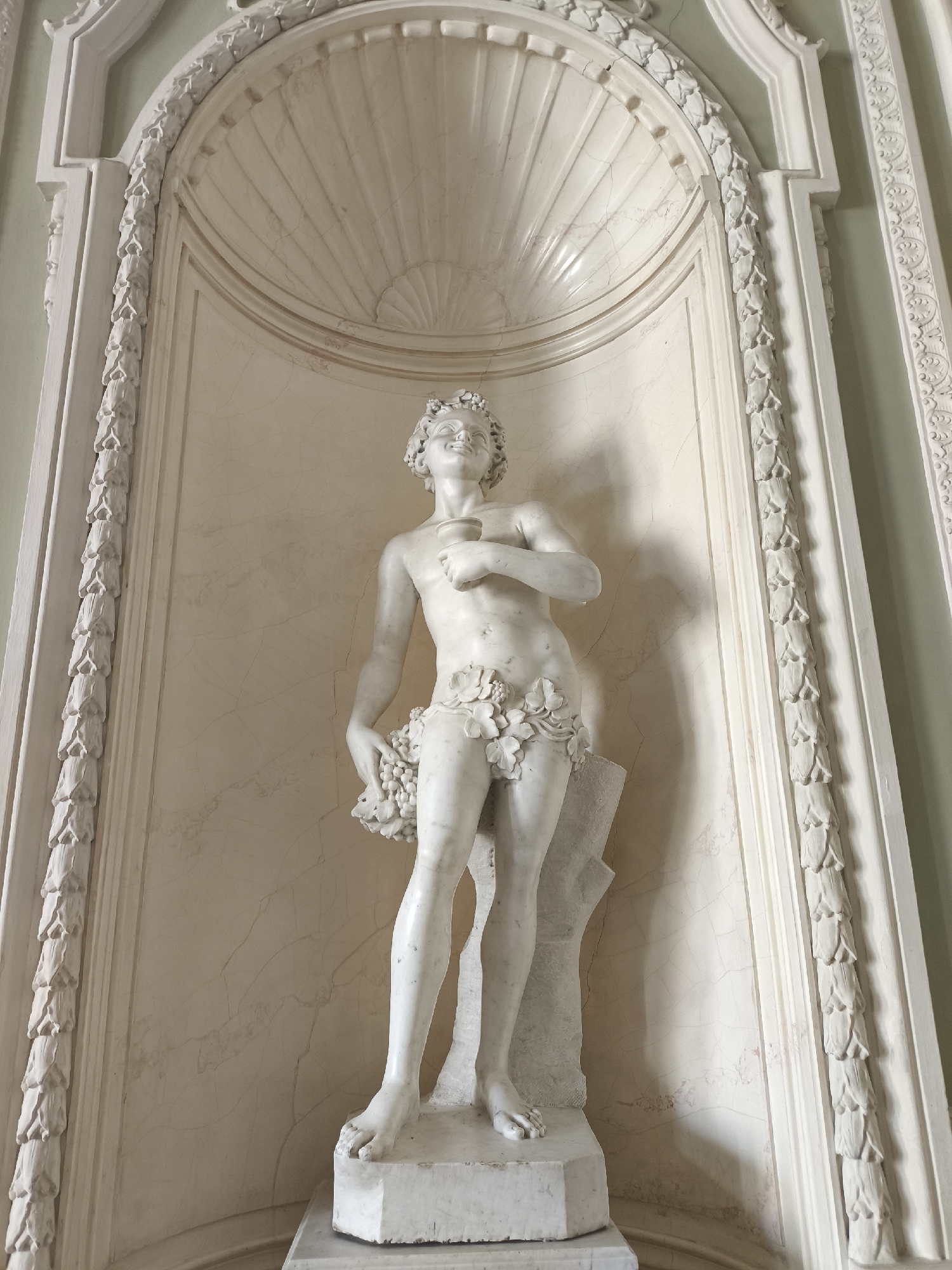 The image shows a white marble statue of a young man, possibly Bacchus or a similar mythological figure, standing in a niche. The figure is depicted with curly hair and a wreath of leaves on his head, a smiling expression, and is partially draped in a garland of leaves and grapes around his waist. He is holding a chalice in his right hand and a bunch of grapes in his left. The statue is placed on a pedestal within a decorative alcove. The alcove features fluted details on the ceiling and decorative molding along its sides. The overall setting appears to be an interior space with classical architectural elements. This statue is located in the Ballroom of the Yusupov Palace on the Moika River in Saint Petersburg, Russia.