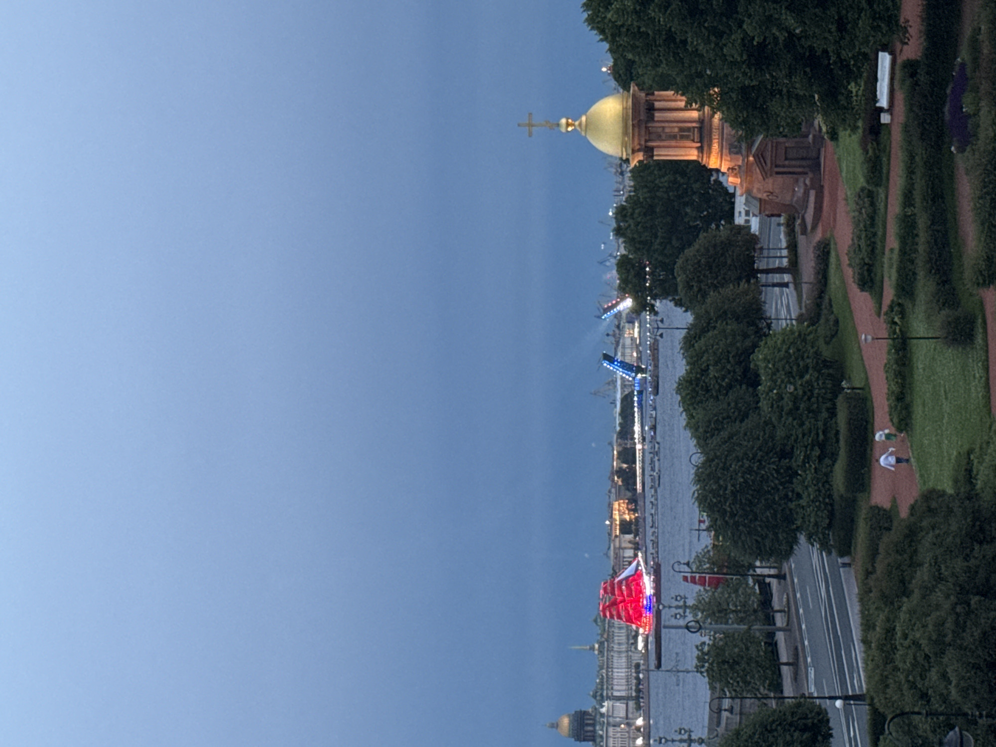 The image depicts a scenic view of Saint Petersburg, Russia, at dusk. The sky is a deep blue, suggesting either twilight or early evening. On the right side of the frame, a well-maintained park area features manicured green hedges, red paved pathways, and mature trees. Two people are visible walking in the park, dressed casually.
Across a wide body of water, the city skyline is illuminated by artificial lights. A notable feature is a bridge, possibly a drawbridge, which appears to be raised or in the process of being raised, with blue and red lights. In the foreground of the cityscape, a ship or boat with a large, red sail, also decorated with lights, is a prominent object. Further in the distance, the golden dome of a building, likely a church or cathedral, is visible, capped with a cross, reflecting the warm glow of the city lights. The overall impression is one of a beautiful urban landscape at a tranquil time of day, with the city lights starting to come alive. There is no visible text in the image.