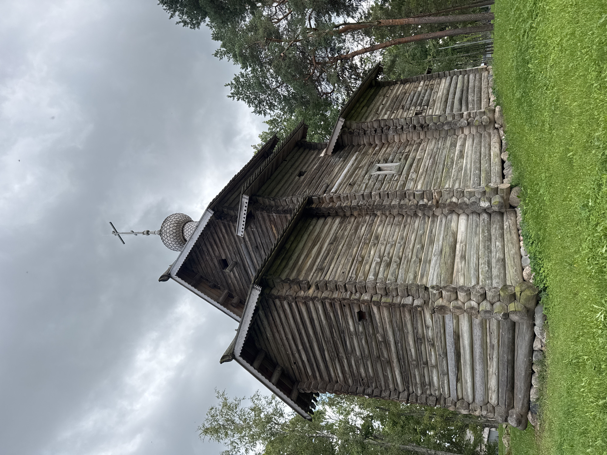 The image depicts a traditional wooden Orthodox church, constructed from logs, situated in Veliky Novgorod, Russia. The church features a distinctive onion dome topped with a cross and intricate wooden carvings along the eaves. The structure is surrounded by lush green grass and tall trees, with a cloudy sky overhead. The architecture suggests a historical or museum setting, possibly an open-air museum or a preserved historical site. No people are visible in the image, and the overall atmosphere is serene and timeless.