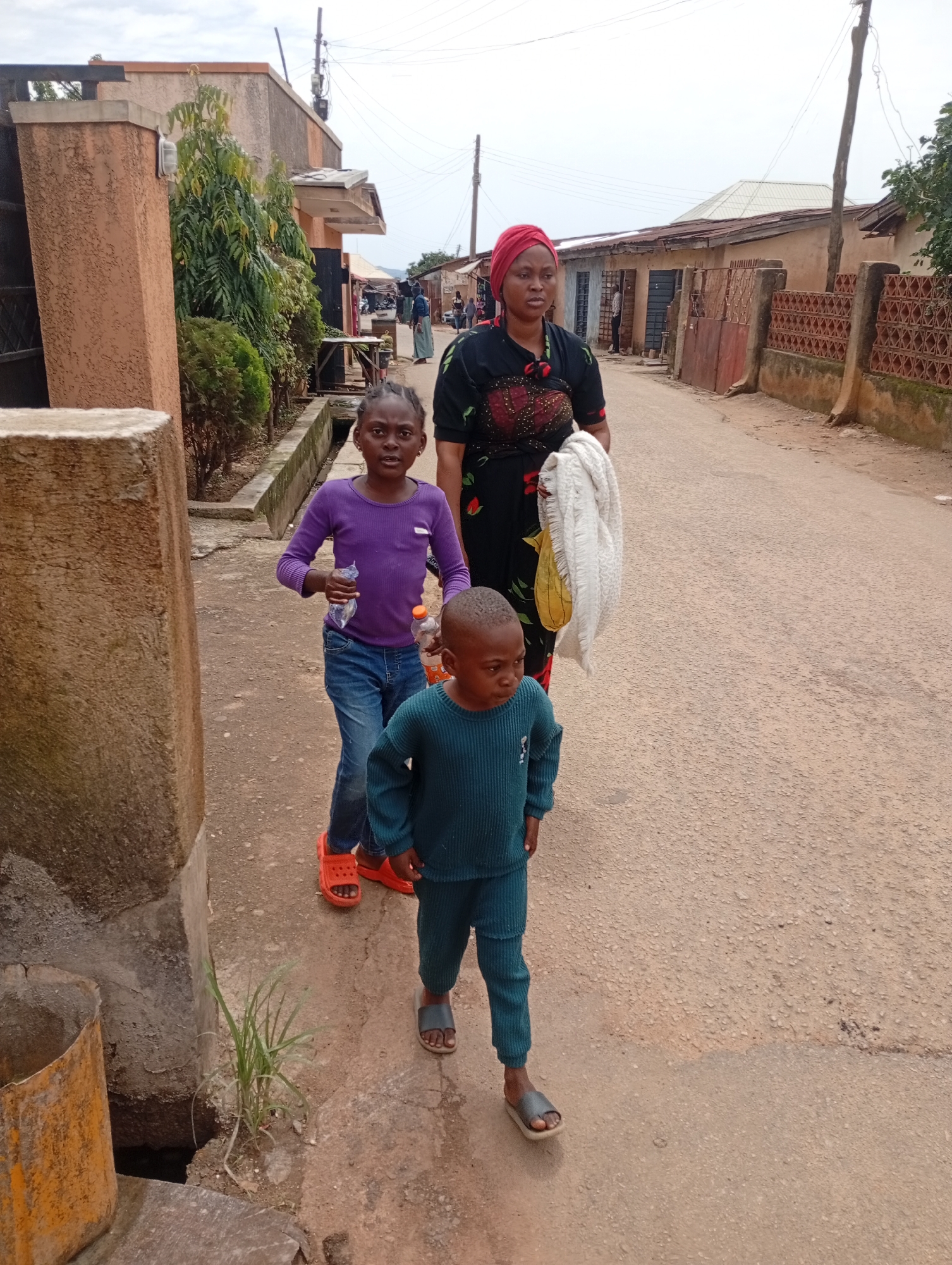In a street scene in Jos, Nigeria, a group of people walk along a paved road. A woman wearing a red headscarf and a black dress with red floral accents is at the forefront, carrying a white cloth over her right arm and a yellow bag in her left hand. To her left, a young girl in a purple long-sleeved shirt and blue jeans walks, holding a clear plastic bottle. A young boy, dressed in a teal-colored sweater and matching pants, walks ahead of the girl. He is wearing dark gray slides on his feet. The road is flanked by buildings and a fence with decorative patterns. Greenery, including trees and shrubs, is visible along the roadside. The sky above is overcast.