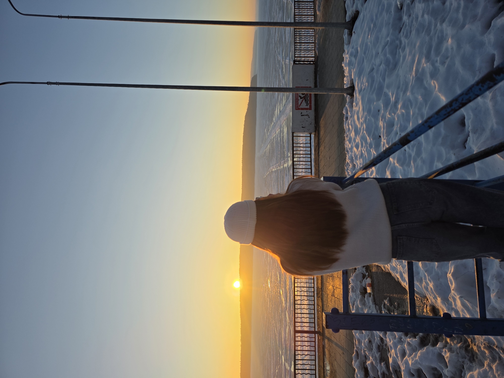 A person stands at a railing overlooking a frozen river at sunset, in the city of Chaikovsky, Russia. The person wears a white hat, white sweater, and light jeans. The railing is blue and the ground is covered in snow. Streetlights are visible in the upper left. The sun is setting over the frozen river.