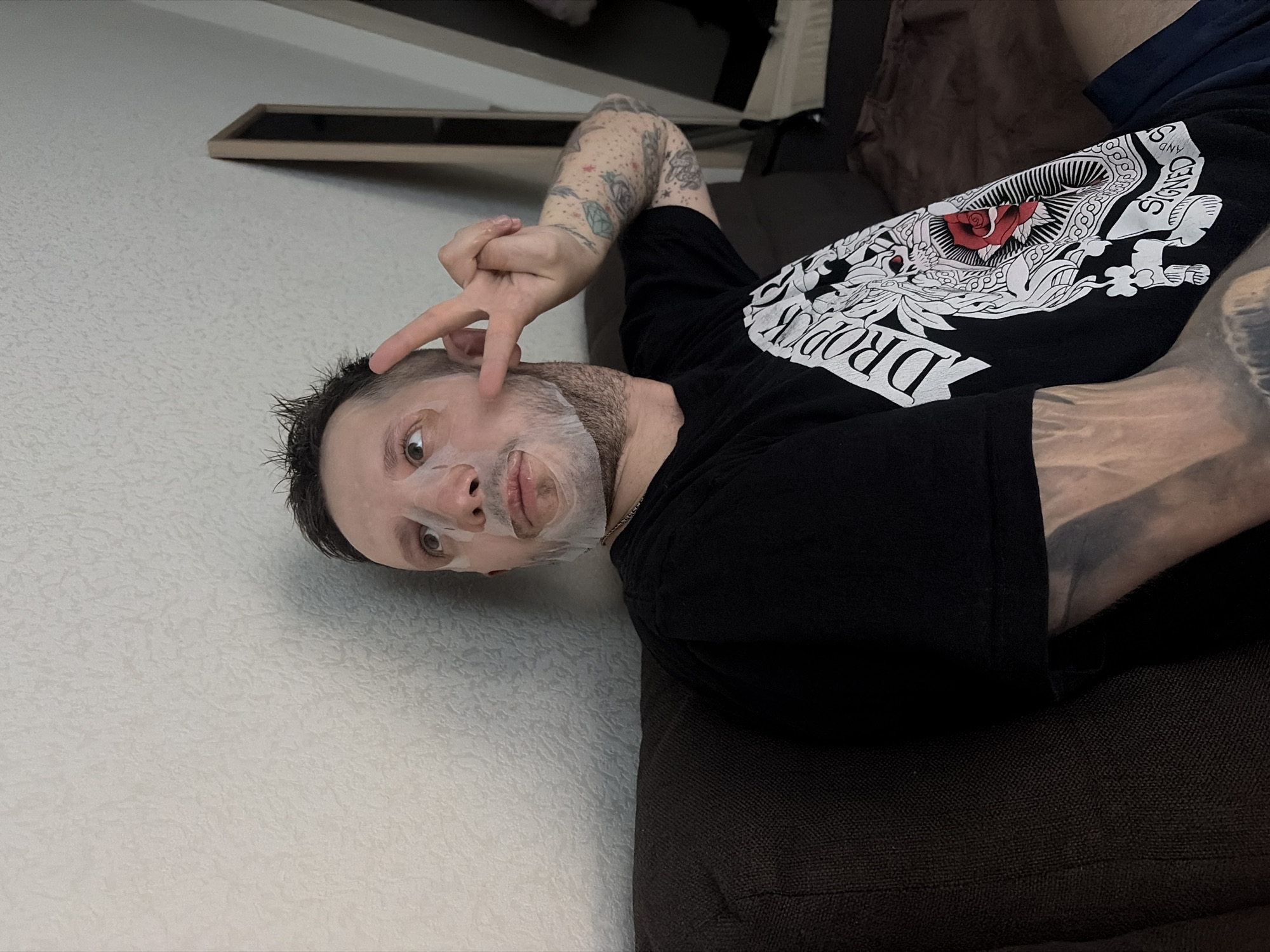 A man lies on a brown couch, wearing a black t-shirt with a graphic design that includes the word "DROPPED" and an image with a red rose. He has a facial mask on and is making a peace sign with his fingers. His arms are tattooed. The background shows a white wall with a rectangular frame-like object mounted on it. The location appears to be indoors, but there is no indication of it being in Saint Petersburg, Russia.