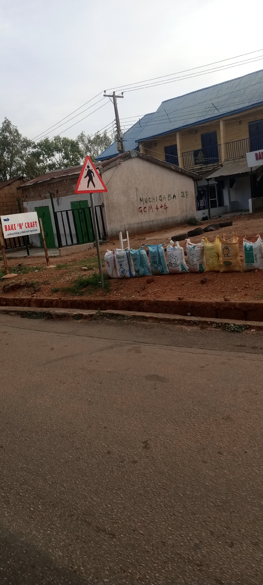 The image shows a street in Jos, Nigeria. A pedestrian crossing sign stands near a "Bake 'N' Craft" sign. Bags of goods are lined up along the curb, and a building with a blue roof is visible in the background. Graffiti is on a nearby wall, reading "MUCHIGABA 27 GCM 4+4".