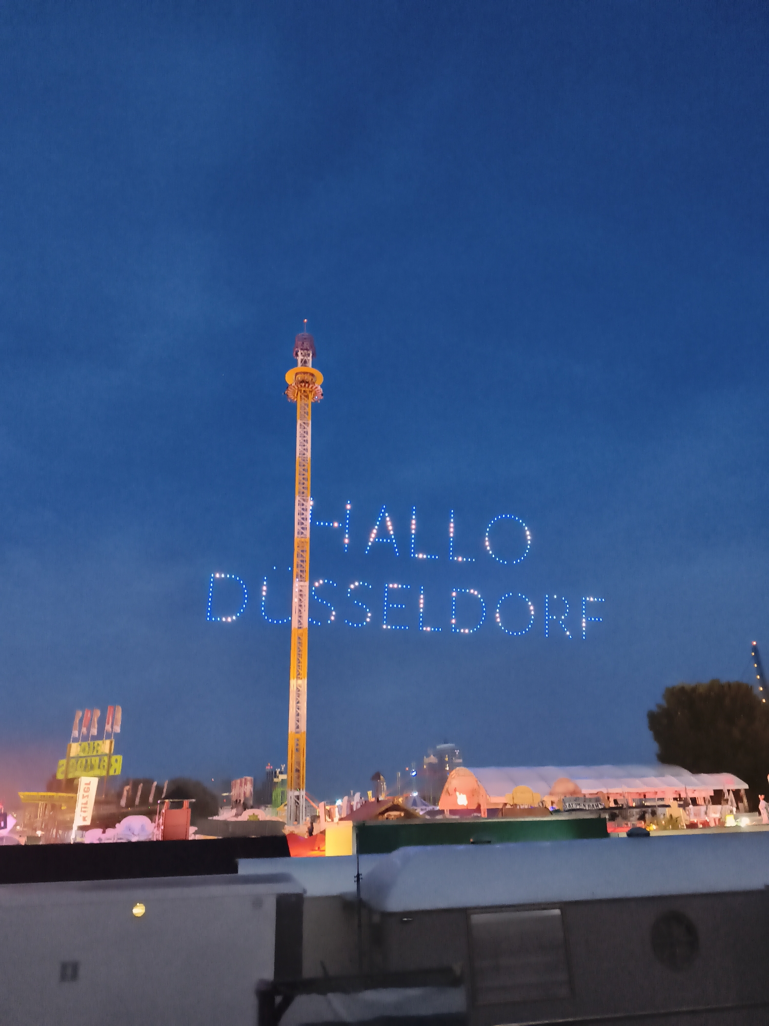 The image shows a nighttime scene at a fairground in Dusseldorf, Germany. Dominating the sky is a tall amusement ride, possibly a drop tower, with bright lights. In the background, illuminated by strings of lights, are various fairground attractions, tents, and structures. The words "HALLO DÜSSELDORF" are spelled out in lights across the dark blue sky, suggesting a festive occasion or welcome message for the city. The foreground features the tops of some buildings or stalls, likely part of the fairground infrastructure. The overall atmosphere is that of a lively outdoor event at dusk or night.