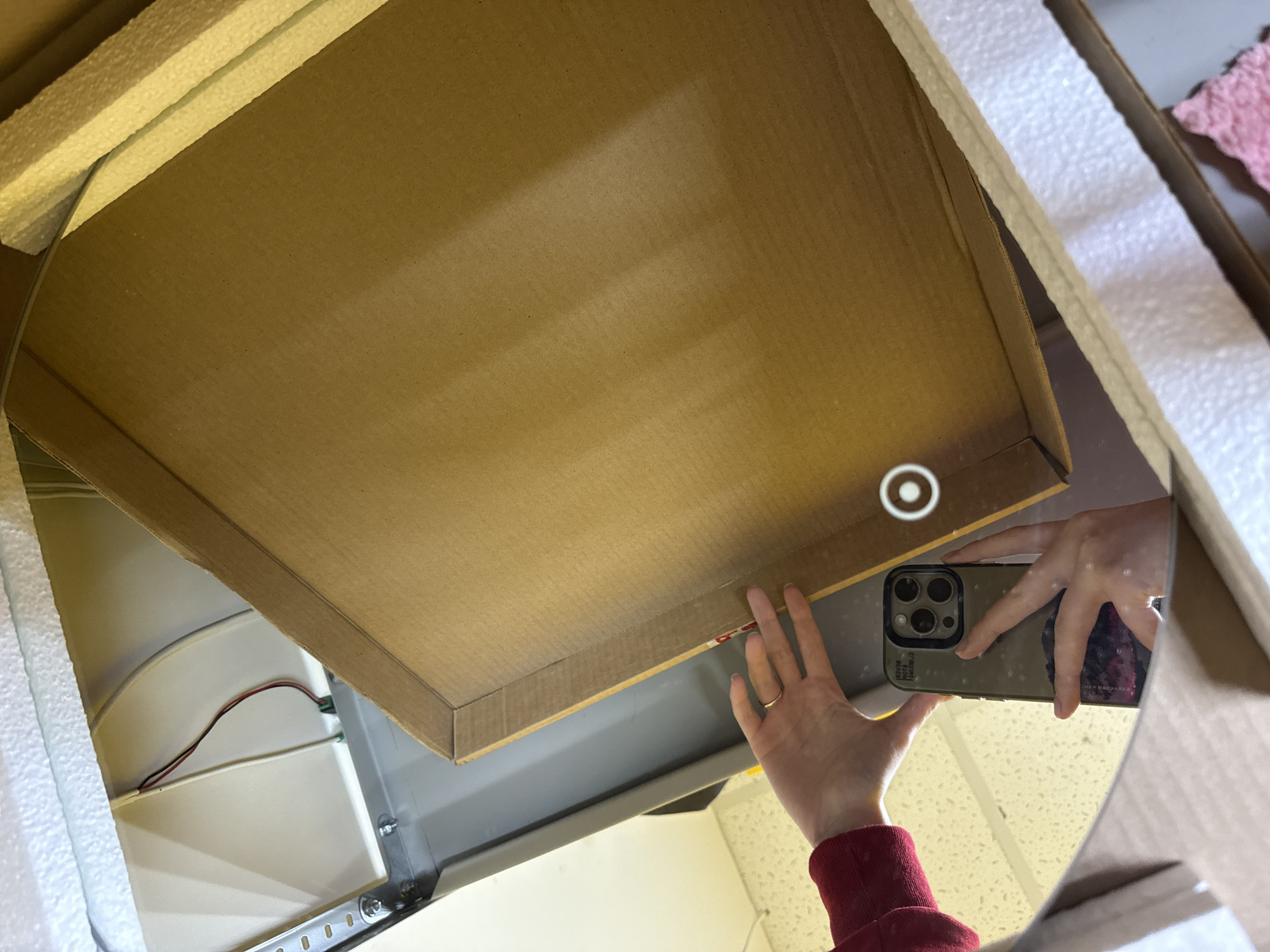 The image appears to be a reflection, likely in a mirror or highly polished surface, capturing a person's hands engaged in unboxing or inspecting an item. One hand holds an iPhone, equipped with a three-camera system and a dark-colored case, positioned as if taking a photo. The other hand reaches into a cardboard box.
Inside the box, white styrofoam packaging material surrounds what seems to be a grey metal frame or appliance, partially revealing internal wiring with red, black, and white cables. The reflected background includes a ceiling featuring a circular recessed light fixture and a light-colored, speckled floor or wall. The person's left arm, visible in the reflection, wears a red sleeve, and a ring is present on one finger of the left hand. Faint, reversed text is visible on the back of the smartphone case, including "DESIGNED BY APPLE IN CALIFORNIA ASSEMBLED IN CHINA" and possibly "CASETiFY" and "MAGSAFE".
