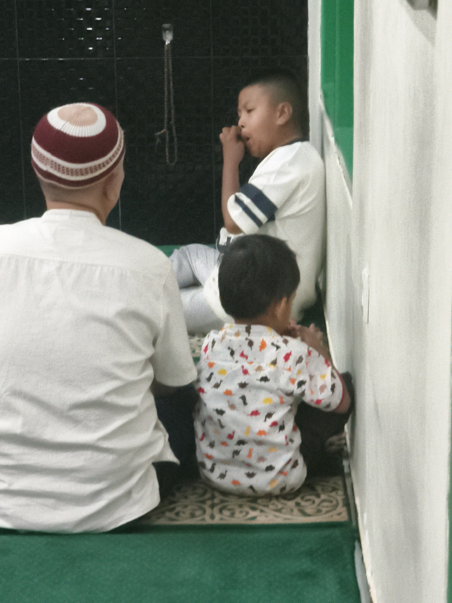 A medium shot captures three individuals, presumably male, inside a building, likely a mosque or prayer room, in Samarinda, Indonesia. The primary focus is on the back of an adult wearing a white shirt and a maroon and white patterned peci (a traditional cap). To the right and slightly in front of the adult, a young boy, facing away from the camera, sits on the floor wearing a white shirt adorned with colorful dinosaur prints. He appears to be engaged in an activity with his hands near his mouth. Further to the right, another boy is visible, leaning against a white wall, also facing away. He wears a white t-shirt with blue stripes on the sleeves and has his hand near his mouth, possibly eating or engaged in a similar action. The floor is covered with a patterned prayer rug, and a green mat is visible in the foreground. The background features a dark, tiled wall with a string of prayer beads hanging from it. The lighting suggests an indoor setting, possibly dim. The architectural elements, such as the patterned wall and the color scheme of green and white, are consistent with religious spaces found in Samarinda.